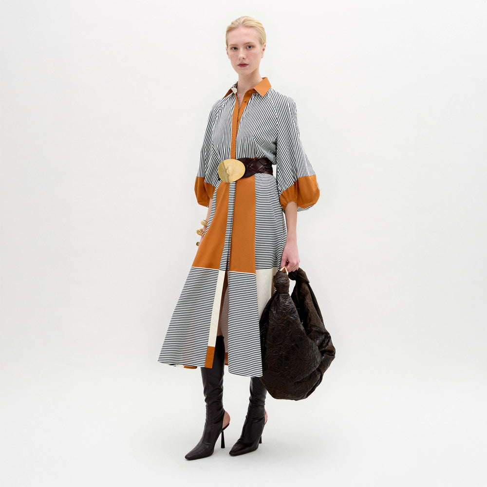 A woman stands against a white background wearing the Agostina Dress—striped and color-blocked, crafted from Italian fabric—paired with high brown boots, a wide belt, and holding a large dark bag.