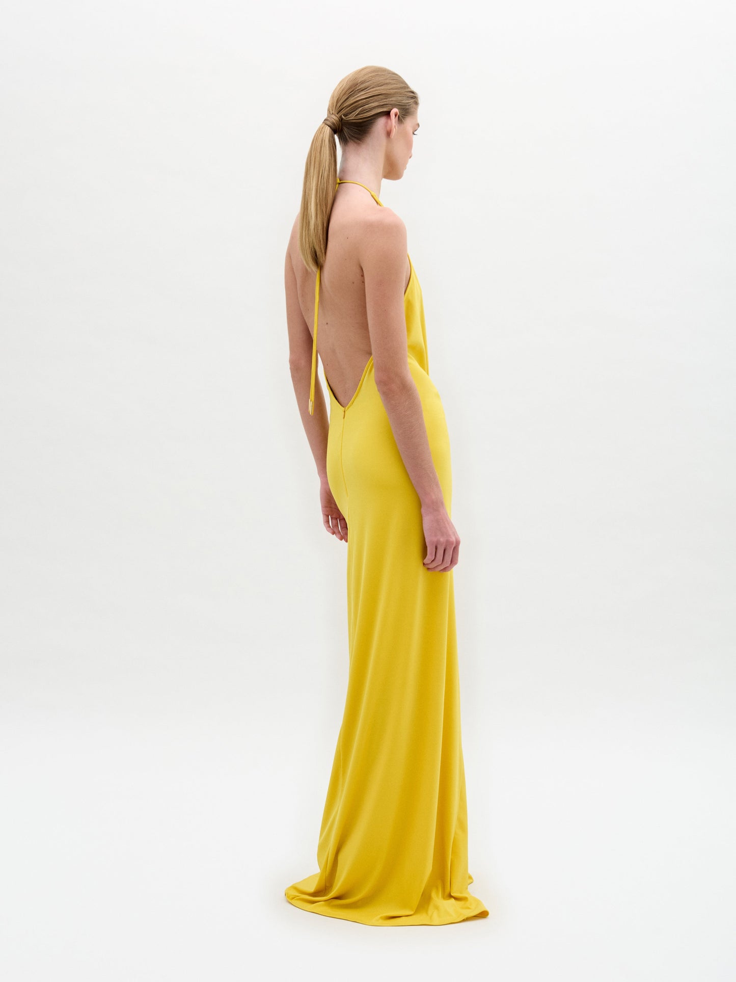 Model in the Aiko Dress—a floor-length, sleeveless yellow maxi with a deep halter neckline and gathered circular waist detail—stands against a plain white background.