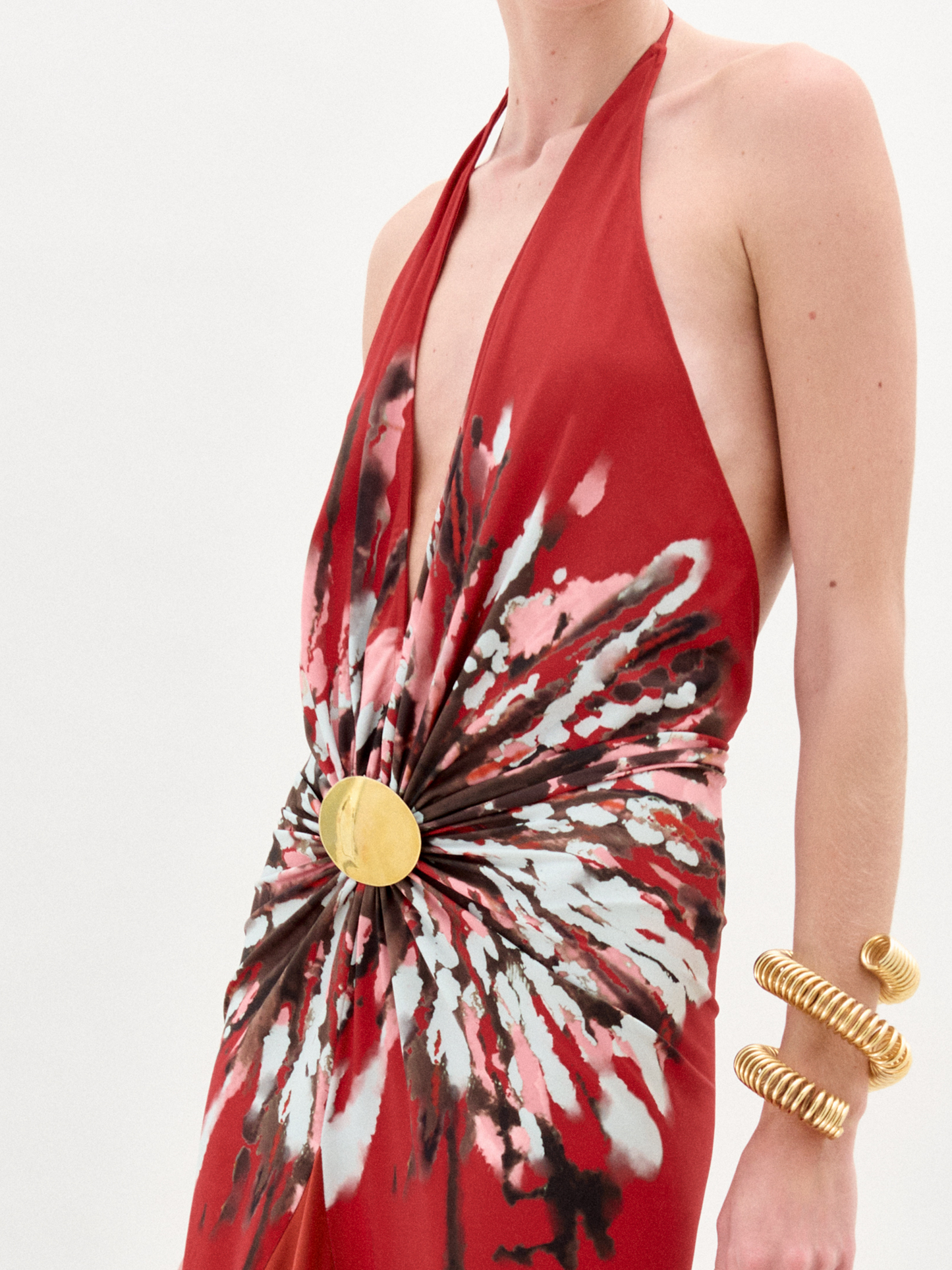 A woman wearing the Aiko Dress—a sleeveless red maxi with a bold white and black burst pattern at the waist and gold embellishments—stands against a plain white background, accessorized with multiple gold bracelets.