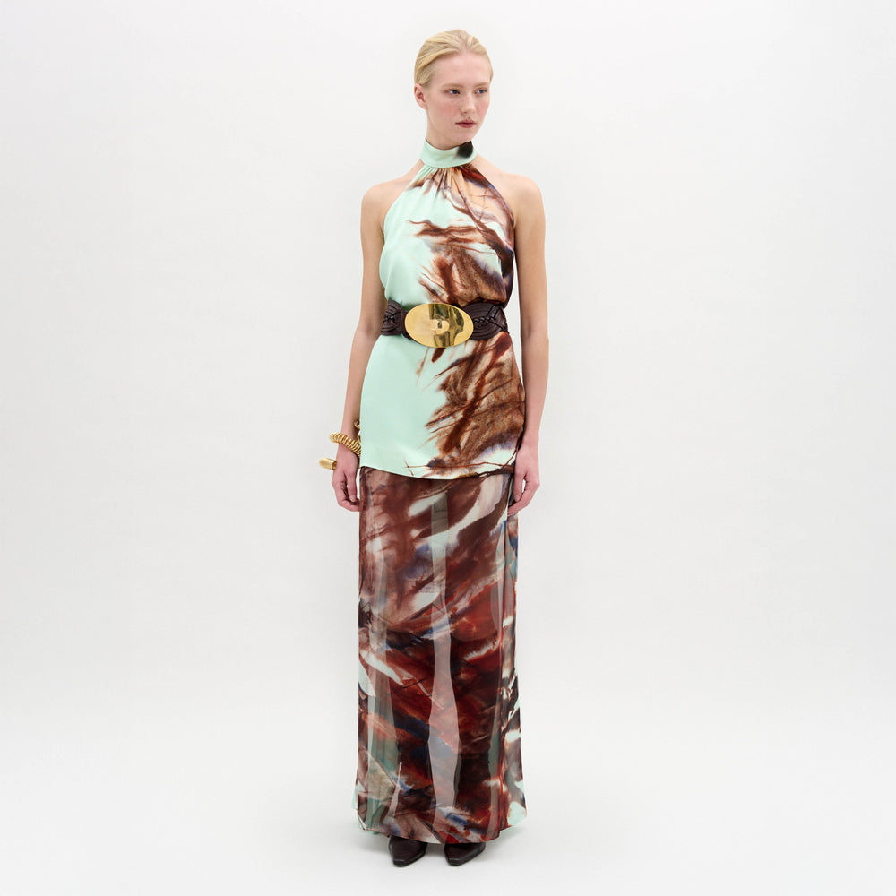 A woman poses against a plain backdrop in the Amalia Skirt—a flowing maxi with an abstract brown and blue pattern—paired with a sleeveless halter top, wide black belt, and bracelets. The look features luxurious Italian fabric.
