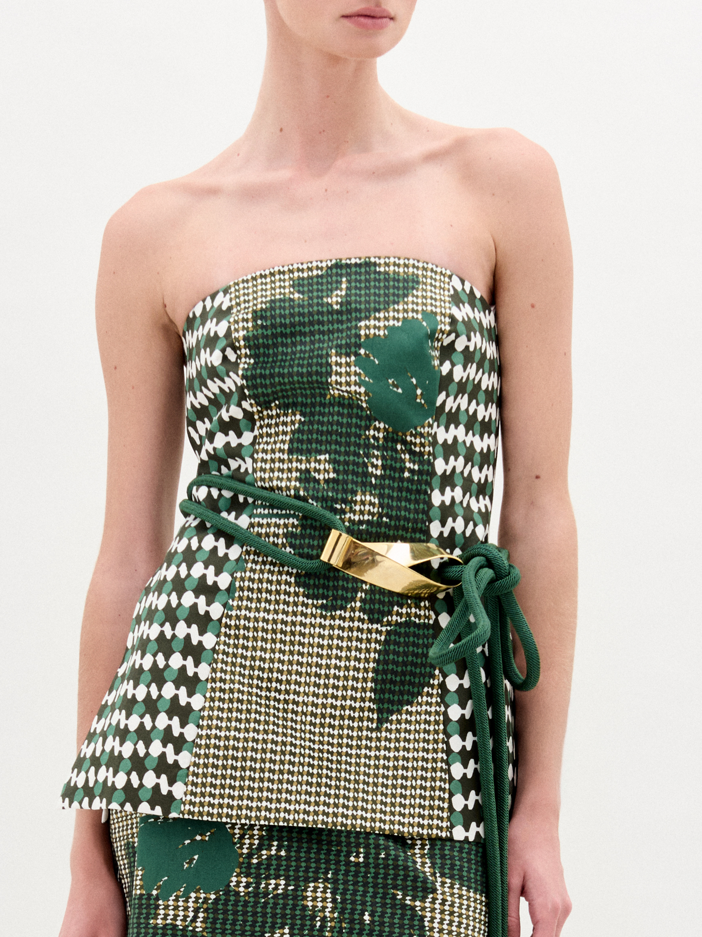 A woman wears the Astor Top, a strapless bustier in green, beige, and houndstooth Italian fabric with a matching skirt and a green belt with gold clasp, standing against a plain white background.