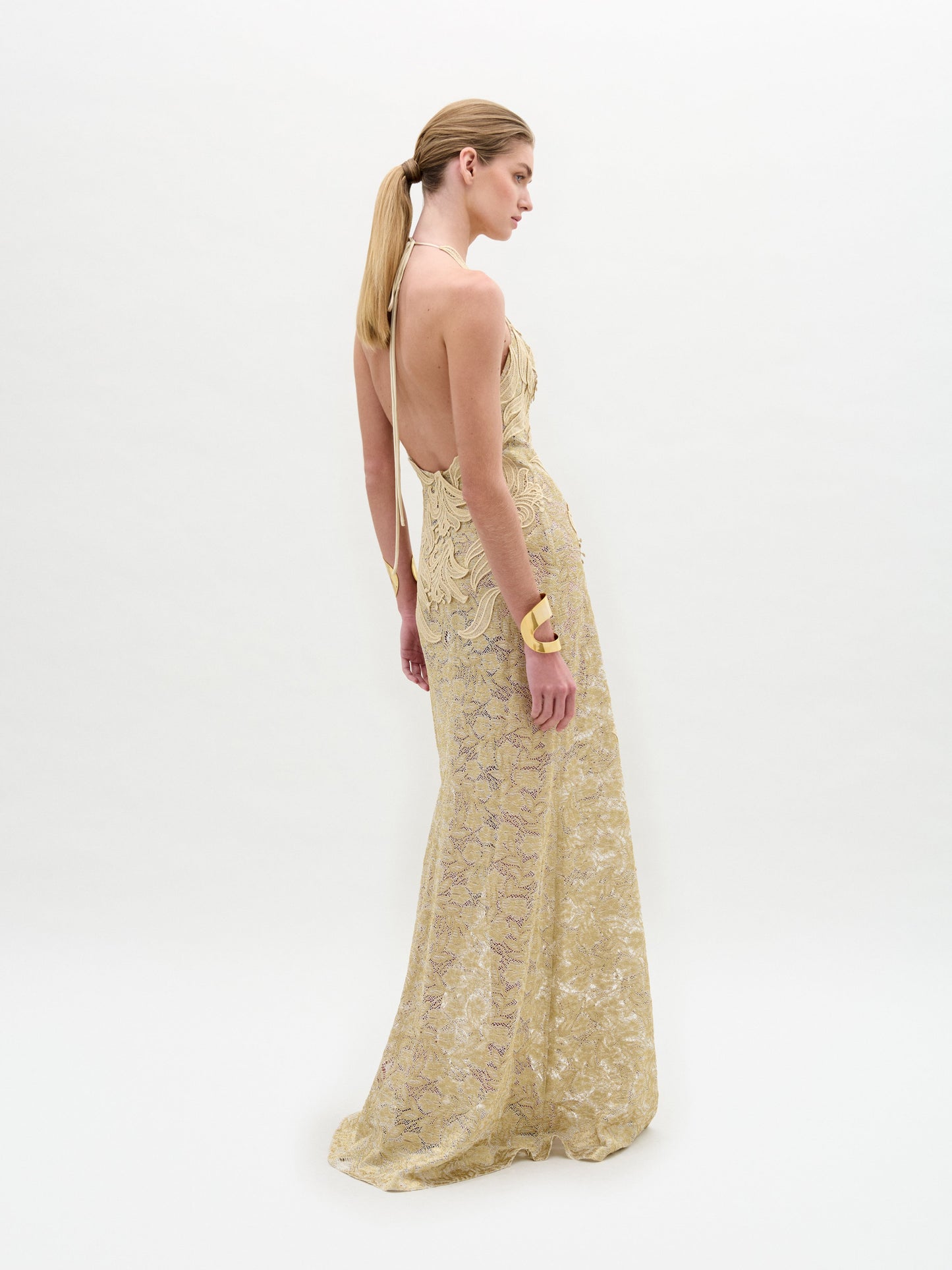 A woman wears the Aurora Dress—floor-length, sleeveless, crafted from gold lace with a fitted silhouette and wavy patterns—paired with gold cuff bracelets, standing against a plain white background.
