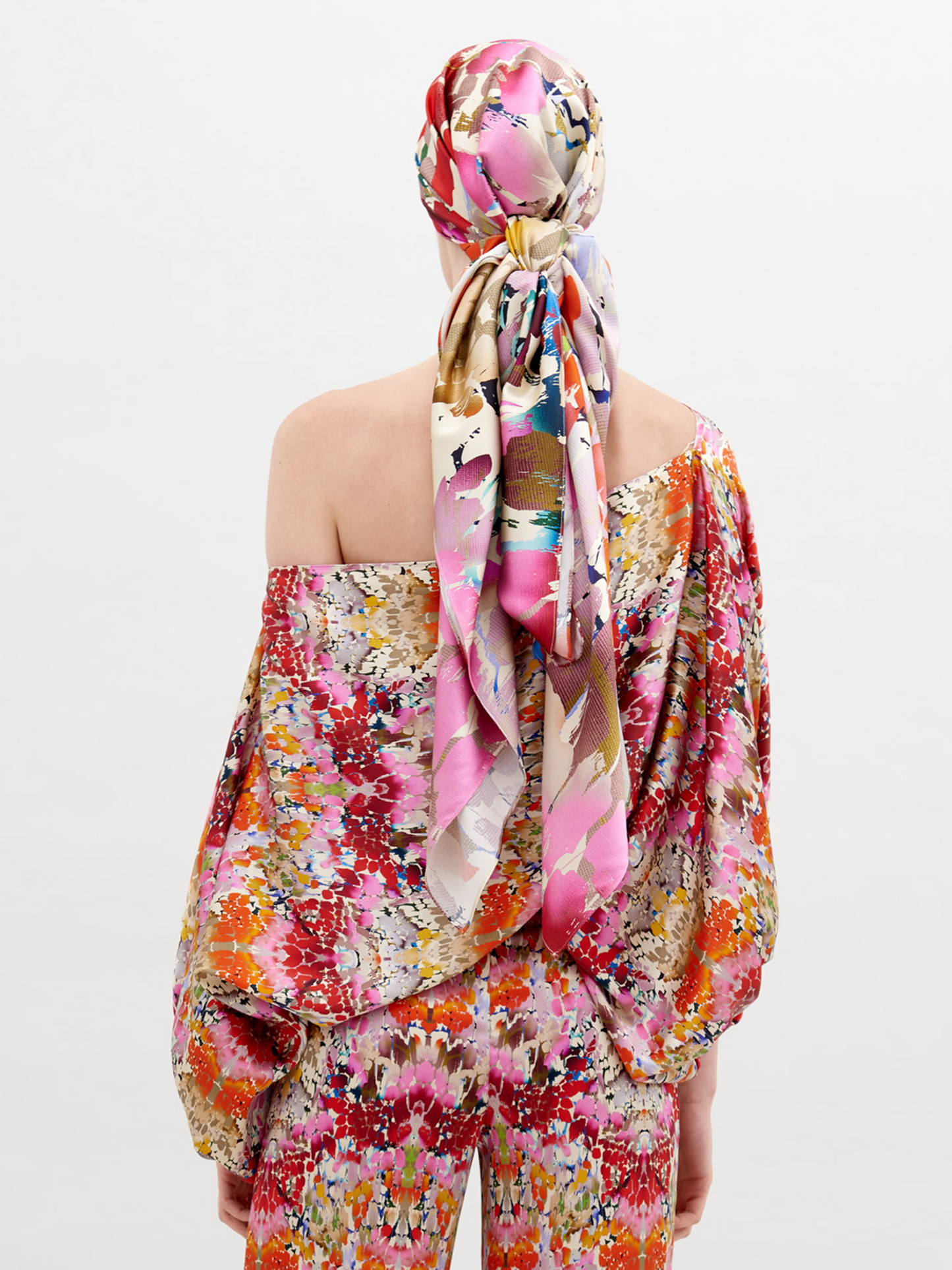 A person stands, wearing a colorful floral outfit with a matching cap. The Bellagio Blouse Multicolor Animal Print has one shoulder exposed, long billowing sleeves, and is paired with matching pants and a knotted belt. Pre-order now to ensure it ships by November 15th, 2024.