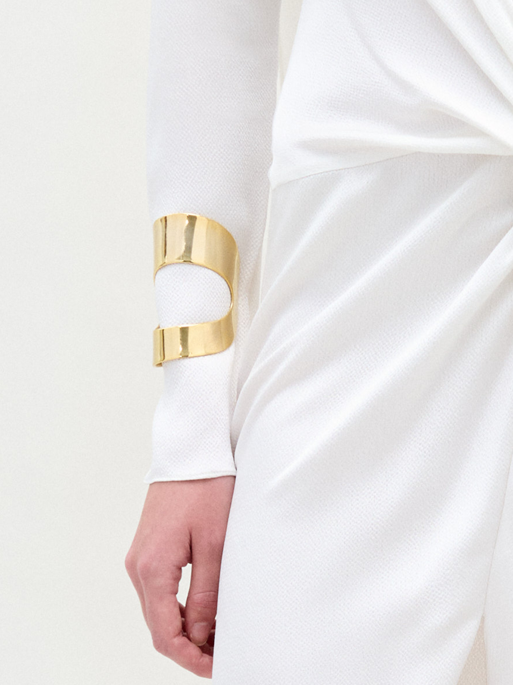 A person wearing a textured white outfit with a gathered waist is shown from the lower arm to hip, showcasing the shiny gold-plated Artemis Cuff on their wrist.