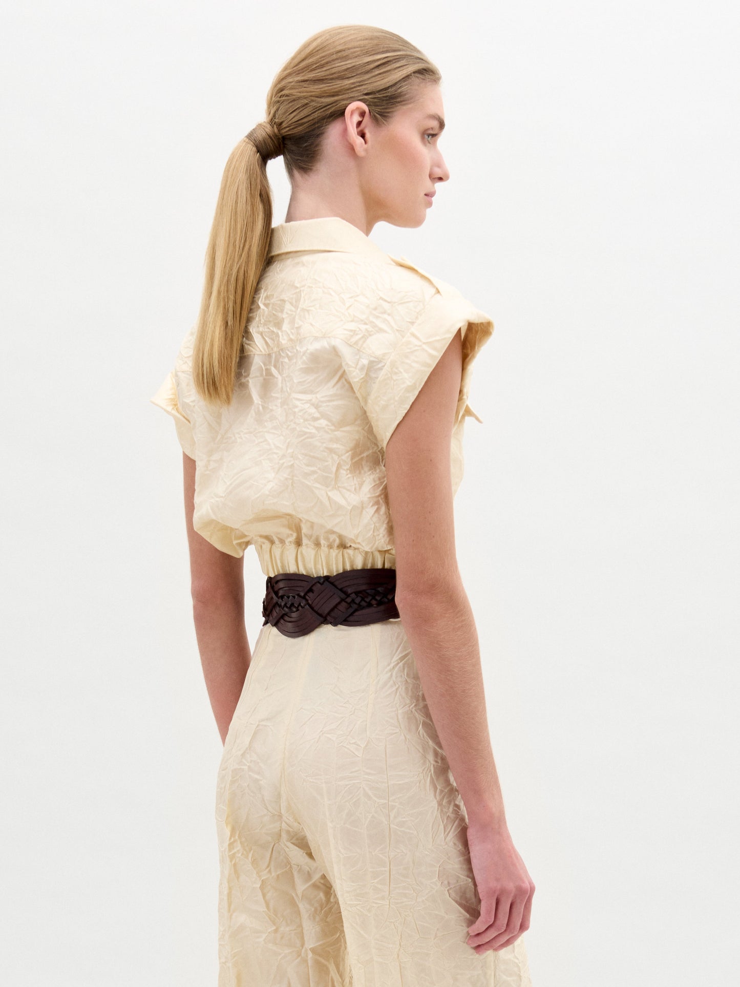 A woman wears the Bara Blouse, a short-sleeved, light beige utility piece made from Italian fabric with front and chest pockets and a wide brown belt with a gold buckle. She stands against a plain white background.