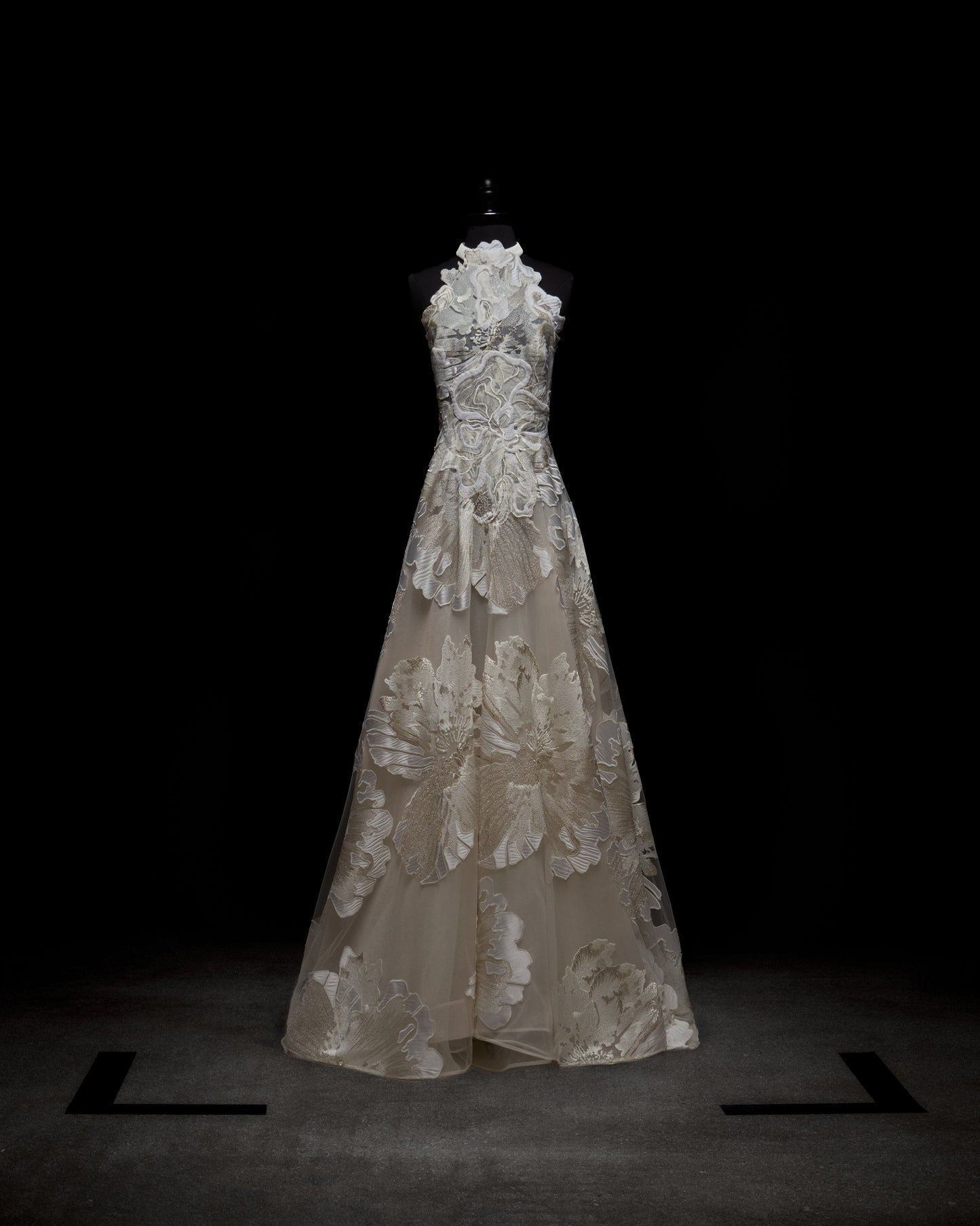 A person wearing the elegant, sleeveless Becca Dress, a white floral gown with a high neckline and sheer gloves, standing against a plain background.