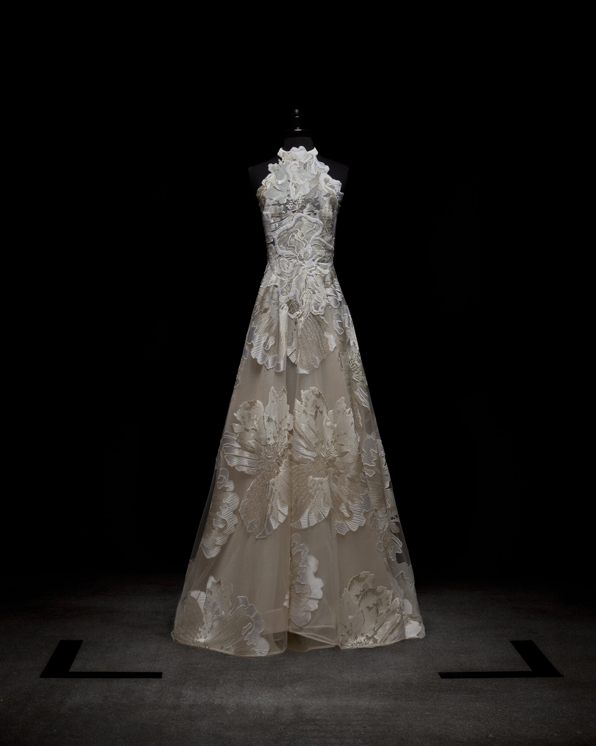 A person wearing the elegant, sleeveless Becca Dress, a white floral gown with a high neckline and sheer gloves, standing against a plain background.