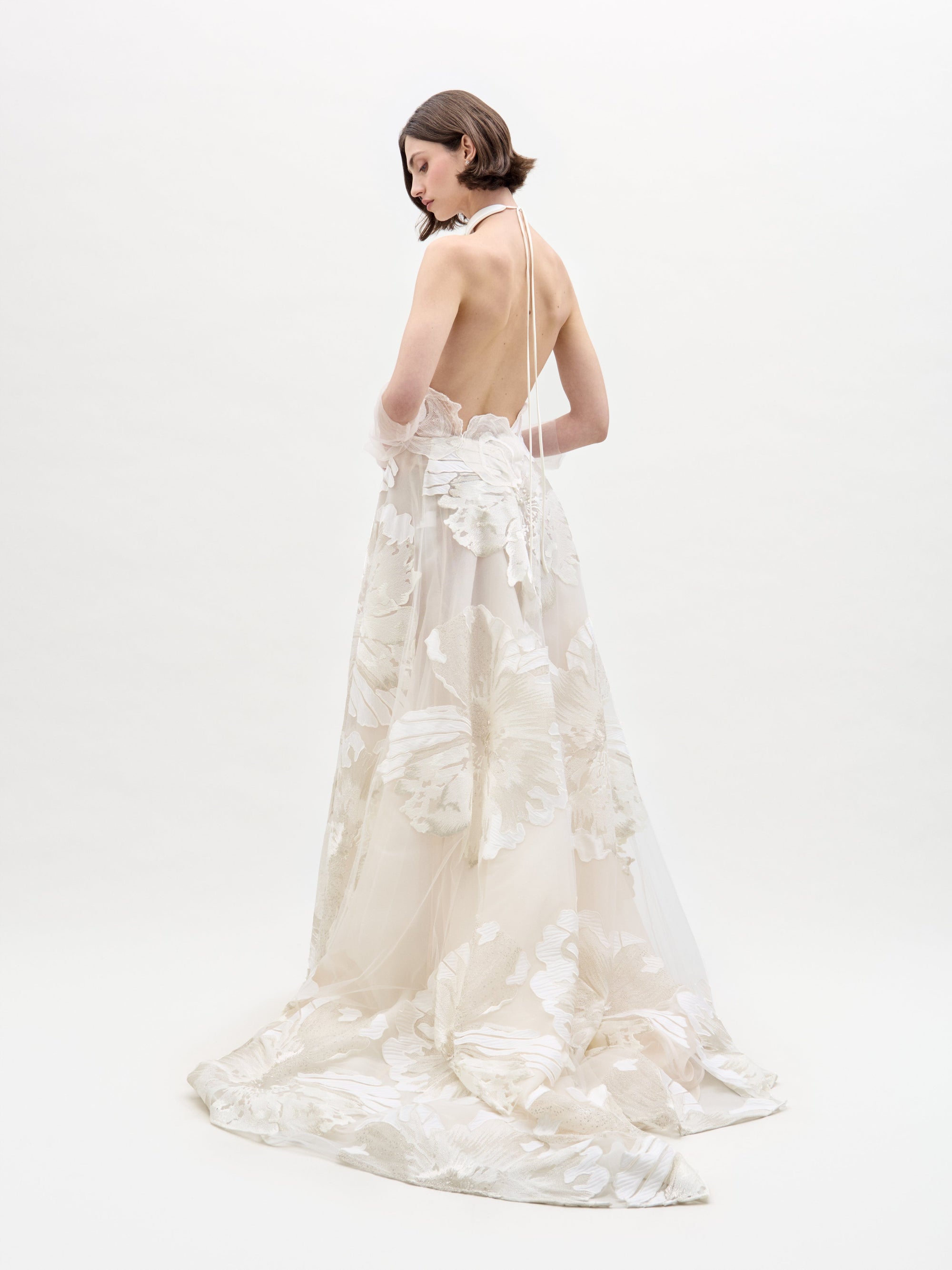 A person wearing the elegant, sleeveless Becca Dress, a white floral gown with a high neckline and sheer gloves, standing against a plain background.