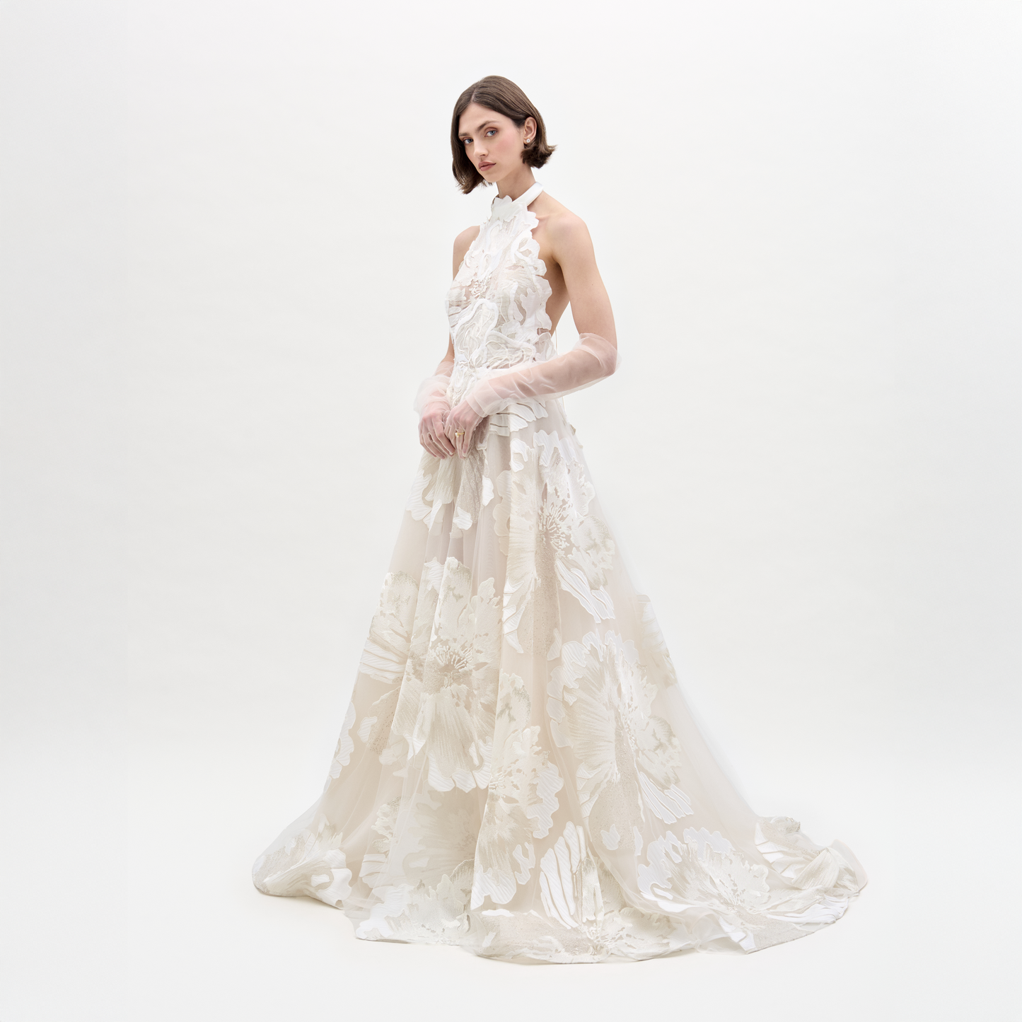 A person wearing the elegant, sleeveless Becca Dress, a white floral gown with a high neckline and sheer gloves, standing against a plain background.