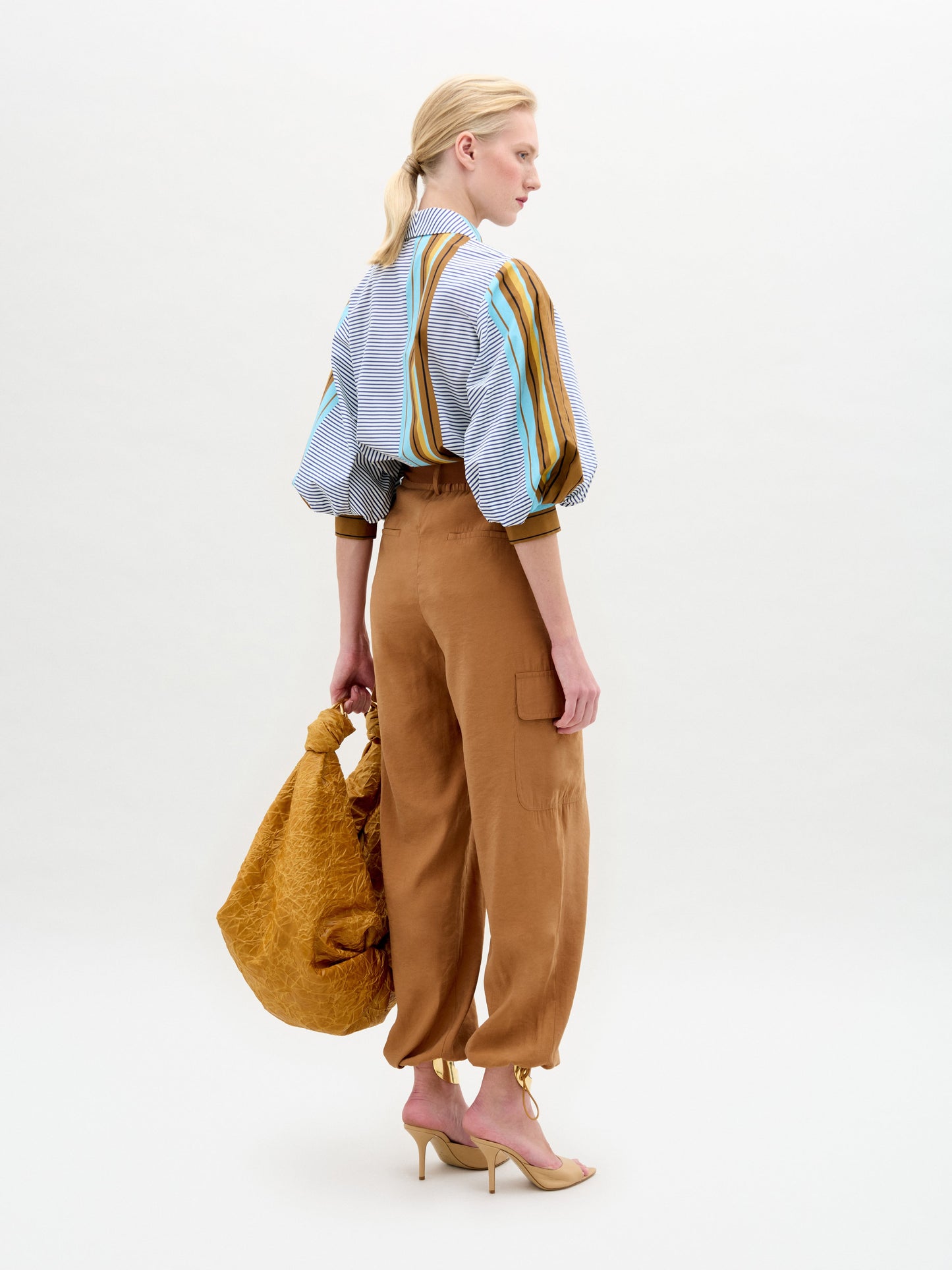 A woman stands against a white background wearing the relaxed fit, high-waisted Darnell Pant in tan cargo style, paired with a striped puff-sleeve blouse, open-toe heels, and holding a large tan bag.