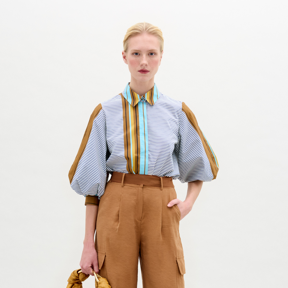 A woman stands against a plain background, wearing the Belkis Blouse with puffed sleeves and oversized tan high-waisted pants, holding a brown bag in her left hand.