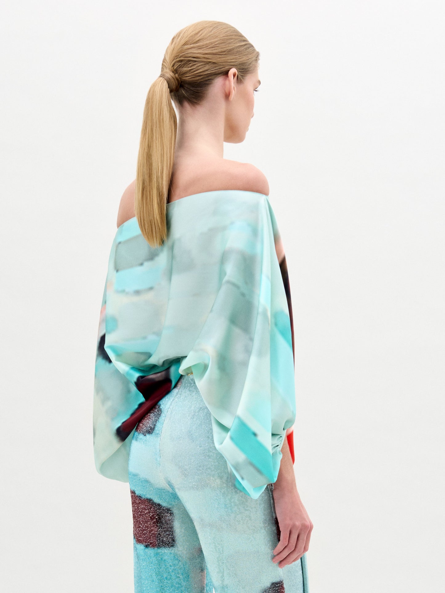 A woman wears the Bellagio Blouse, an off-shoulder red and aqua abstract print silk top with an oversized fit, paired with light blue pants featuring a large circular yellow belt buckle, against a plain background.