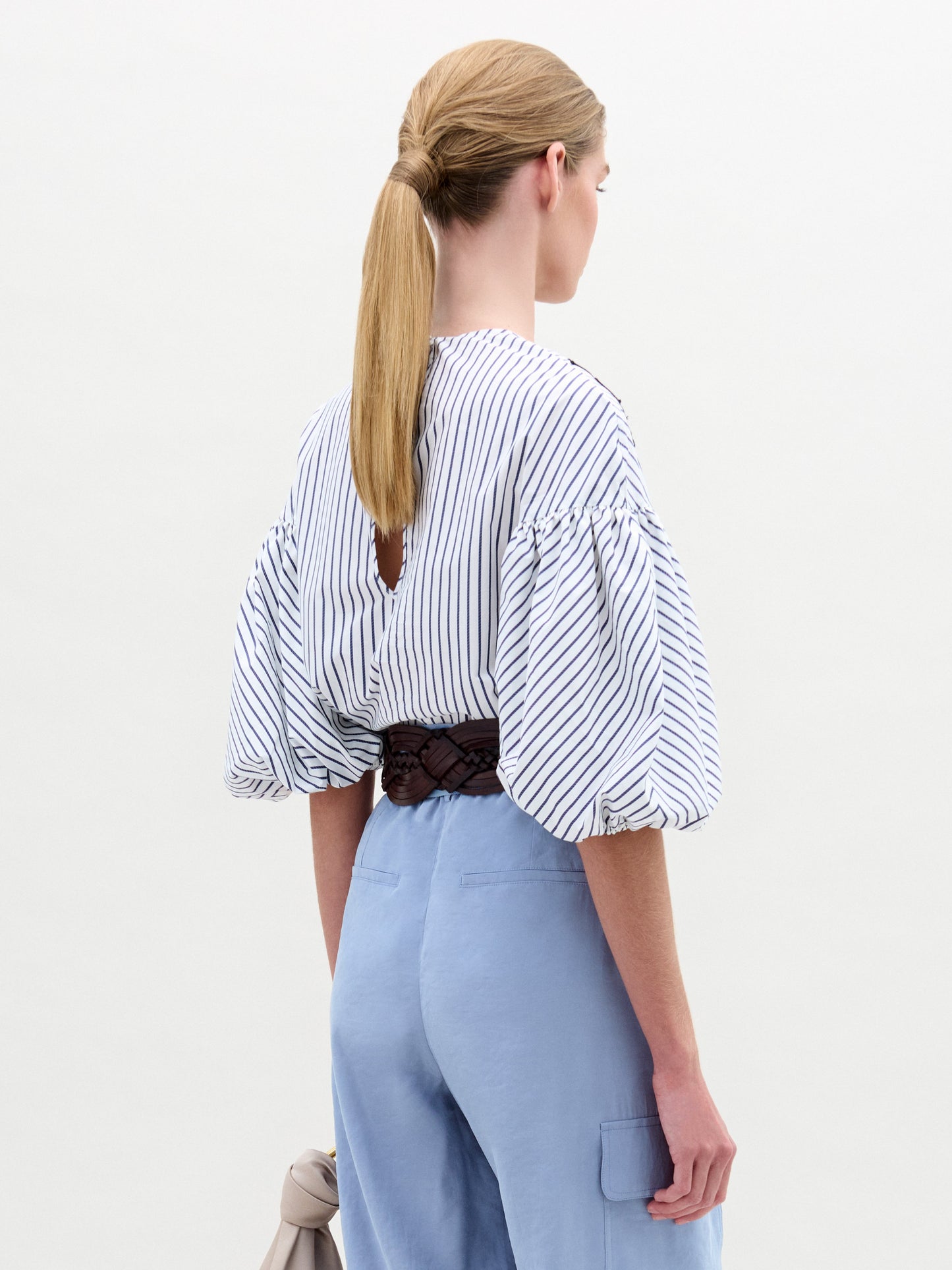 A woman in a Belle Blouse, featuring Italian fabric with stripes and floral embroidery, voluminous sleeves, stands against a plain background wearing light blue pants and holding a small bag.