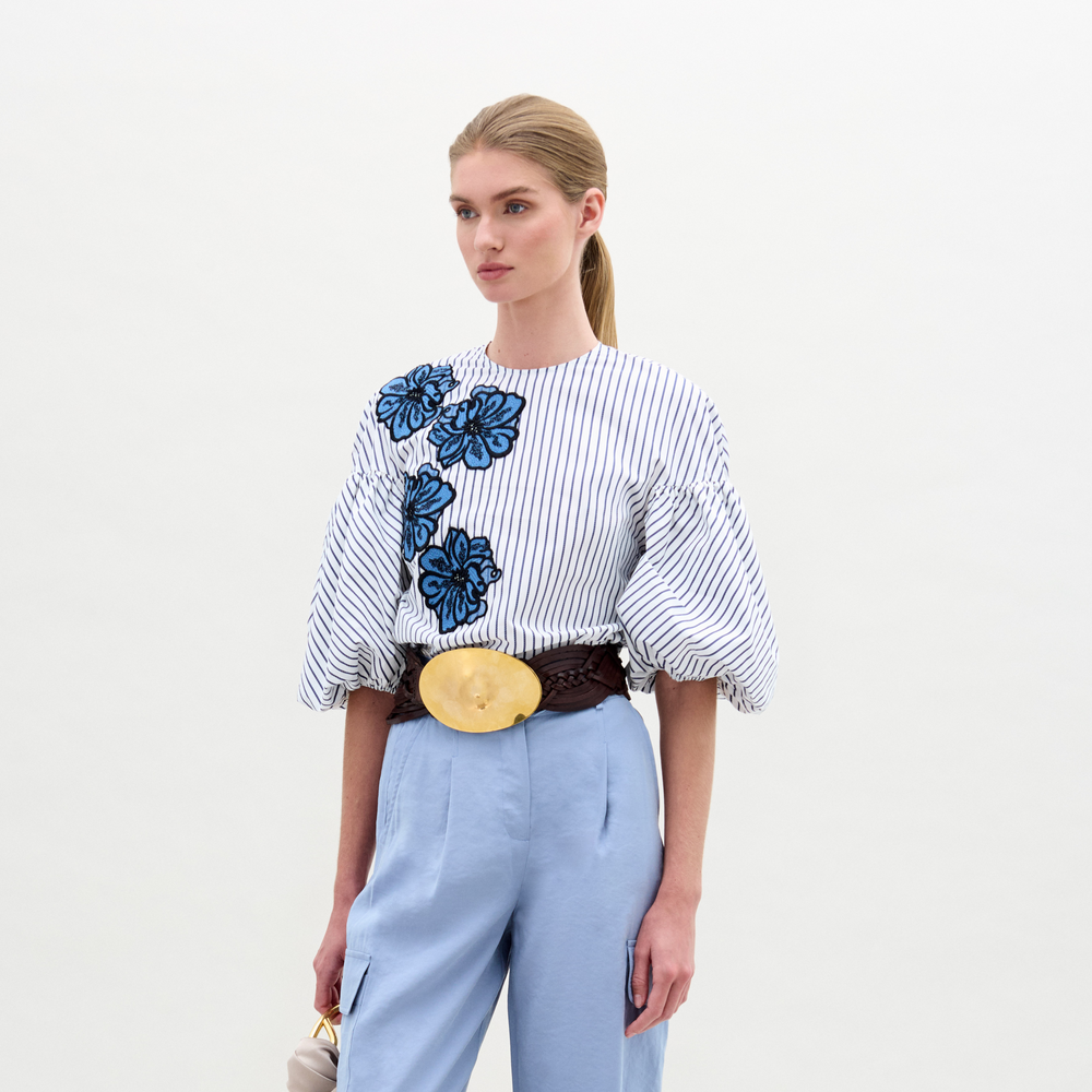 A woman in a Belle Blouse, featuring Italian fabric with stripes and floral embroidery, voluminous sleeves, stands against a plain background wearing light blue pants and holding a small bag.
