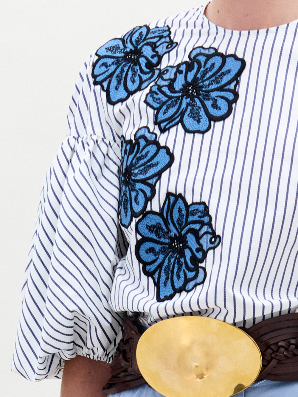 A woman in a Belle Blouse, featuring Italian fabric with stripes and floral embroidery, voluminous sleeves, stands against a plain background wearing light blue pants and holding a small bag.