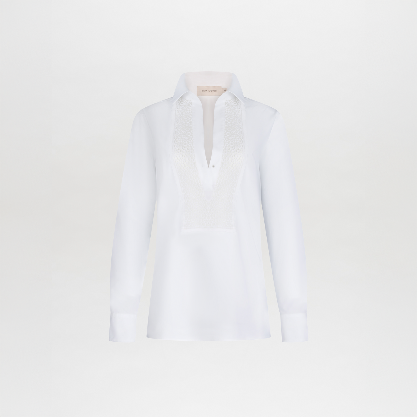 The Bega Blouse is a white, long-sleeve cotton top with a collar and textured lace detailing on the chest, shown against a plain white background.