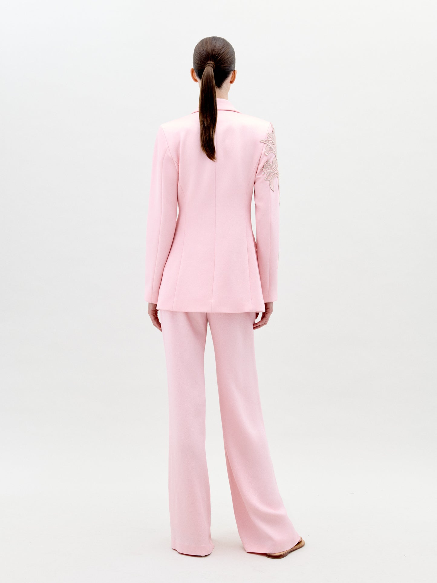 A person stands wearing the Palermo Pant Pink pantsuit, featuring floral detailing on the left shoulder and sleeve, against a plain white background. Available for pre-order now, this elegant outfit will have an exclusive ship-by date of February 1st, 2025.
