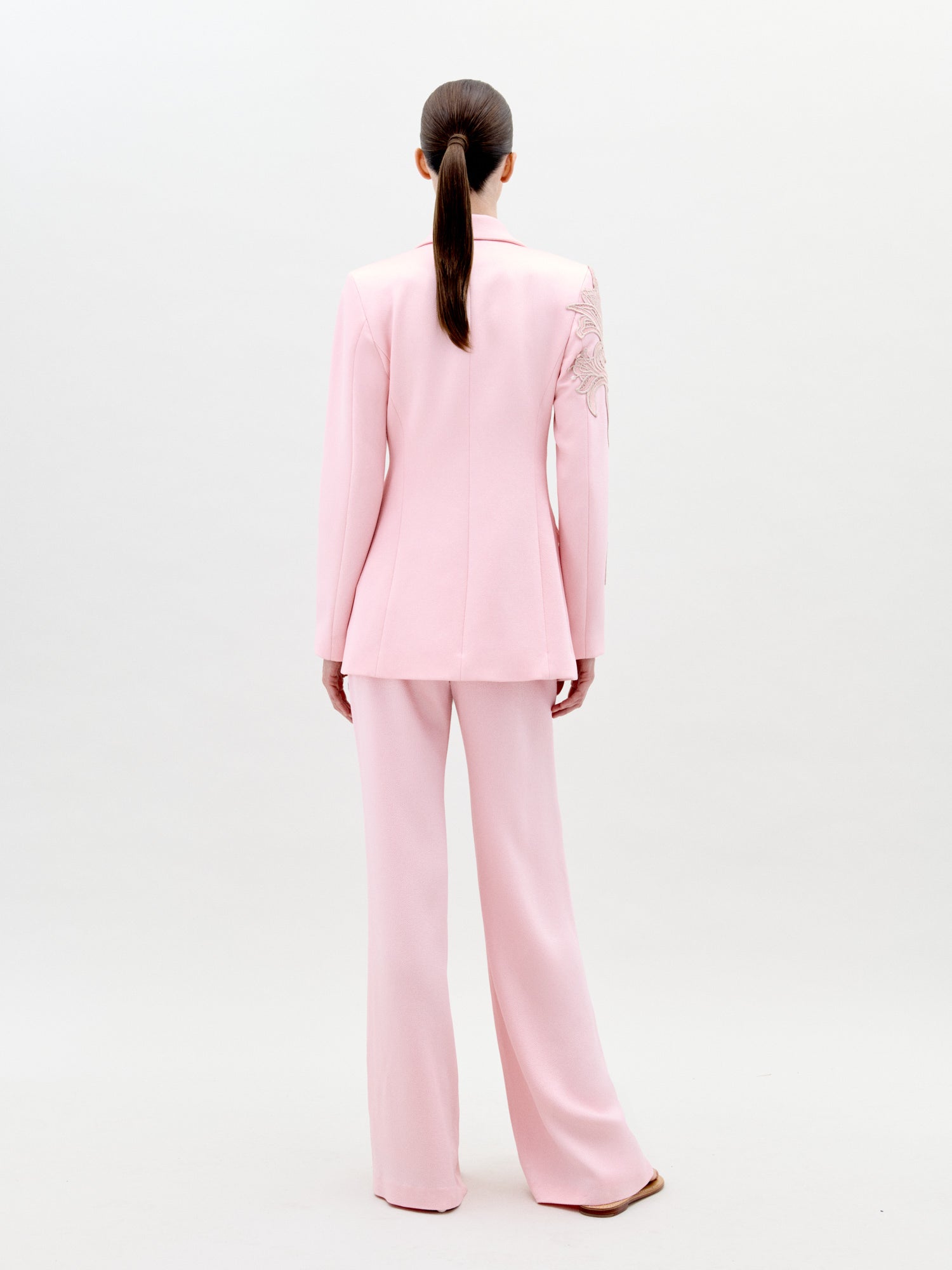 A person stands wearing the Palermo Pant Pink pantsuit, featuring floral detailing on the left shoulder and sleeve, against a plain white background. Available for pre-order now, this elegant outfit will have an exclusive ship-by date of February 1st, 2025.