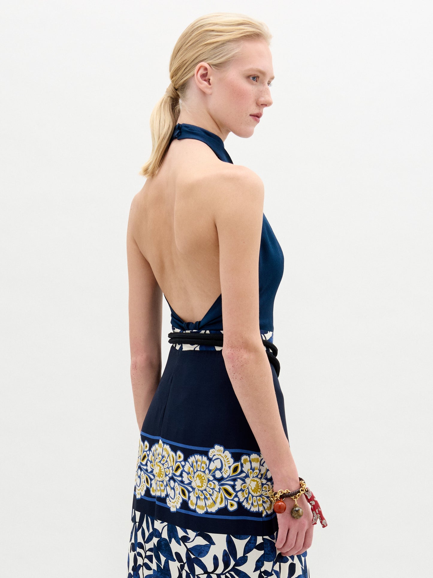 A blonde woman models the Colima Top, a blue high neck crop top, paired with a patterned skirt and large gold belt buckle against a plain white background. This chic outfit is made in Colombia.