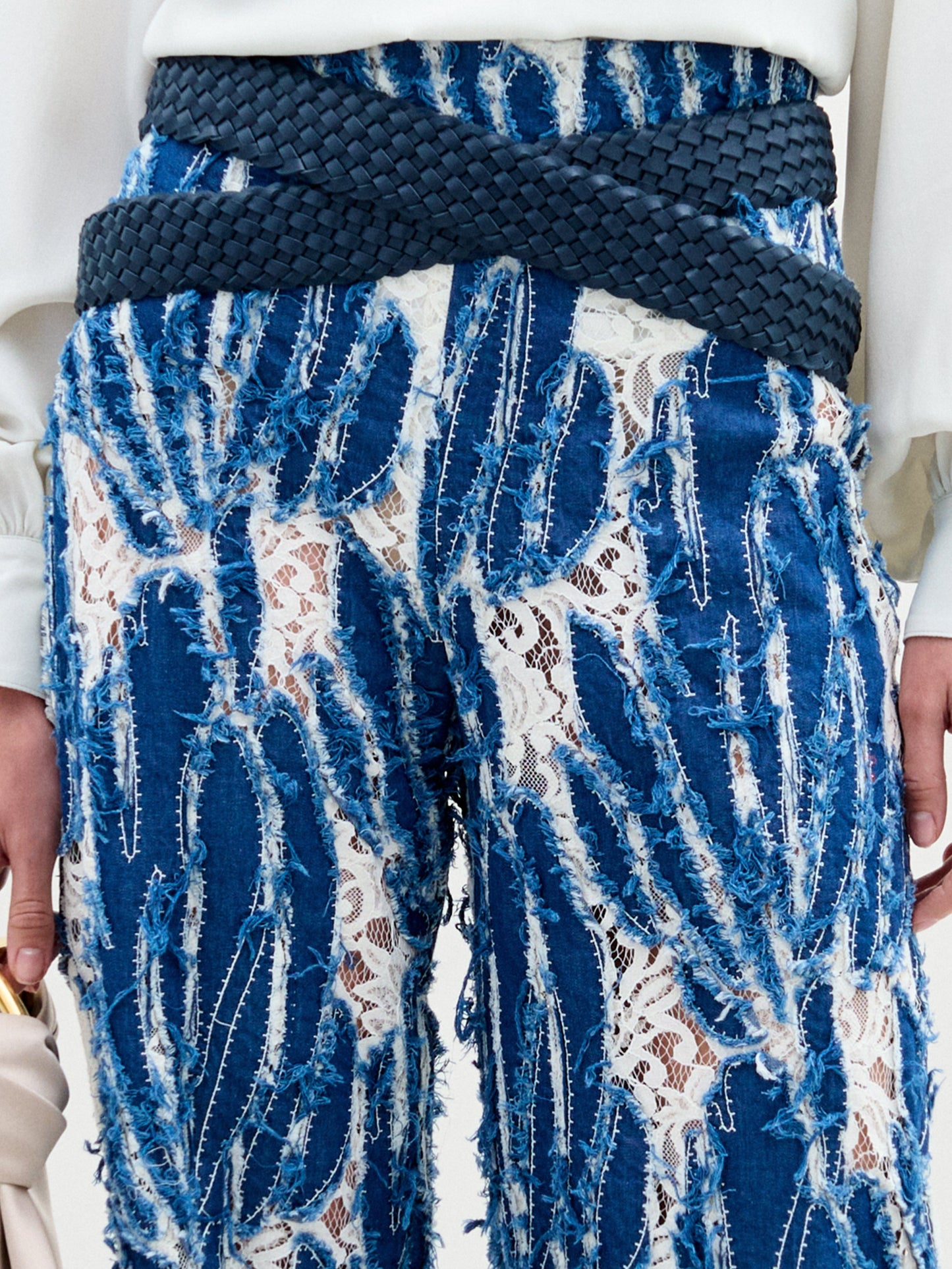 A woman wears the Como Pant, a pair of blue patterned wide-leg pants with a relaxed silhouette, styled with a white long-sleeve front slit blouse and holding a large beige bag against a plain background.