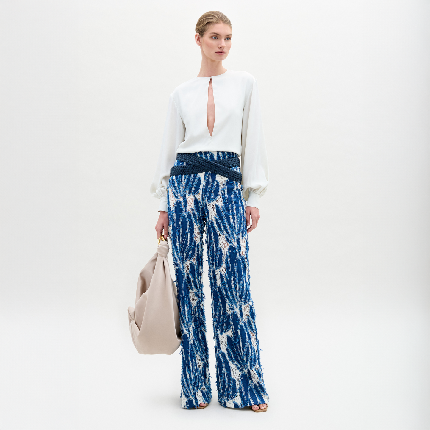 A woman wears the Como Pant, a pair of blue patterned wide-leg pants with a relaxed silhouette, styled with a white long-sleeve front slit blouse and holding a large beige bag against a plain background.