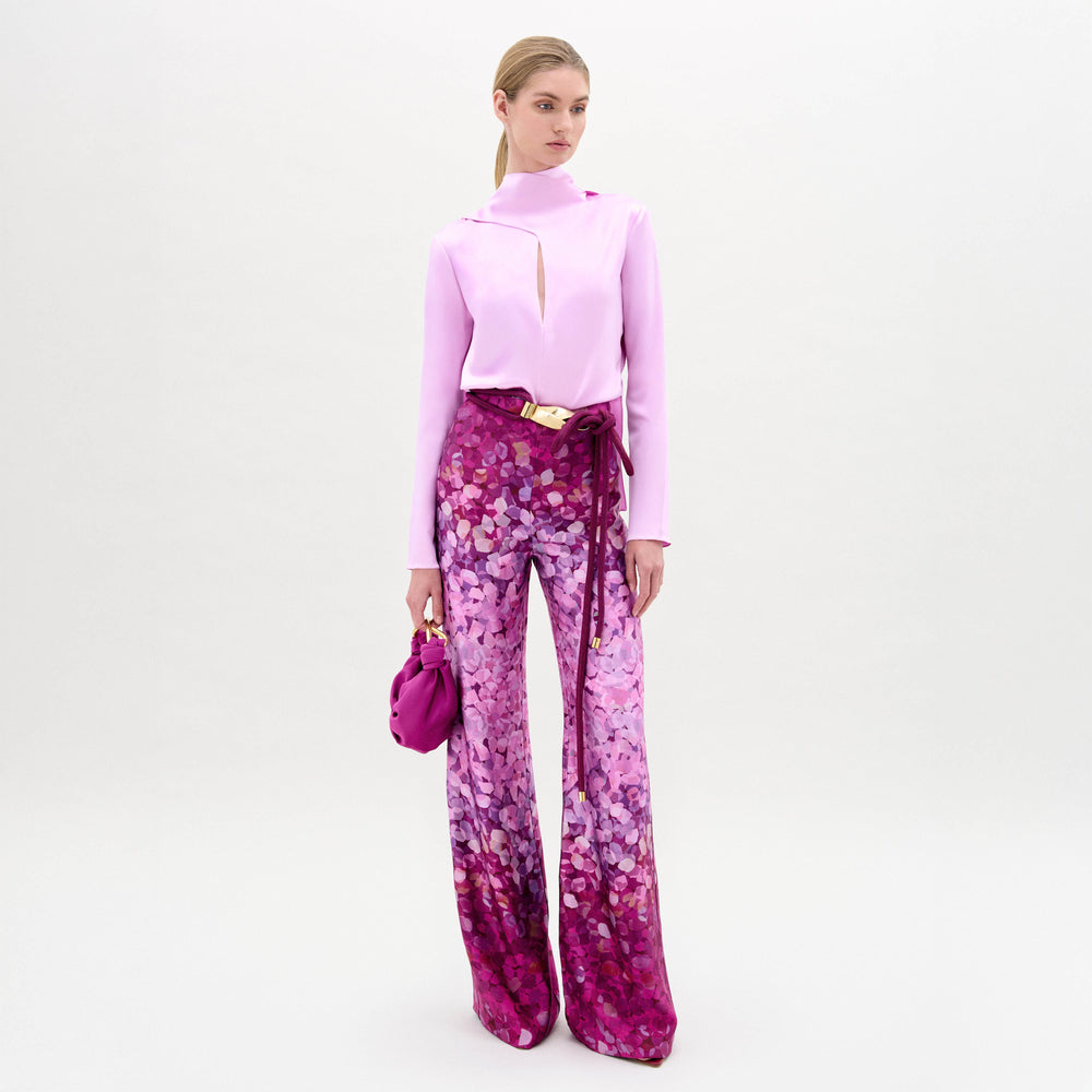 A woman stands facing forward, wearing a light pink long-sleeve top, Como Pant in high-waisted Italian fabric with pink and purple patterns, and holding a matching pink handbag.