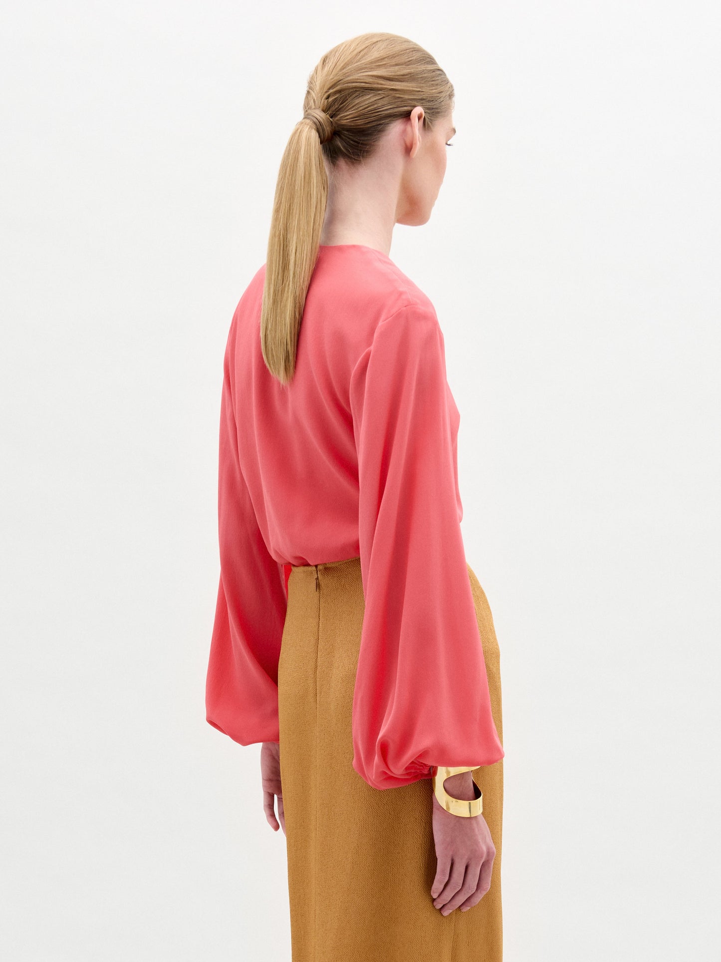 A woman stands against a plain background wearing the Covilha Blouse, a silk coral long-sleeve top, paired with a tan skirt featuring a large gold circular belt detail.