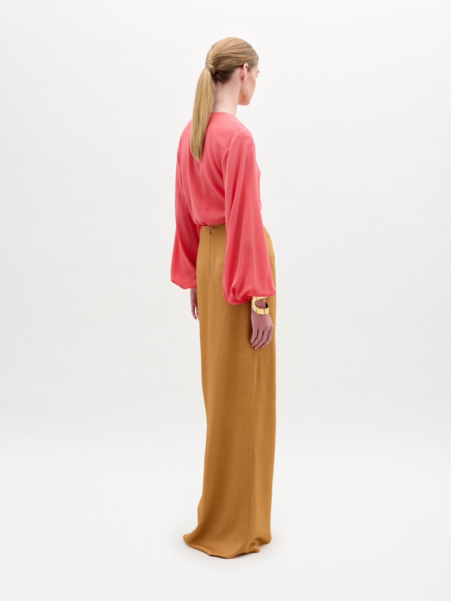 A woman stands wearing a long-sleeve coral blouse tucked into the Gadea Skirt, a high-waisted hammered satin maxi with a striking gold-tone embellishment at the waist.