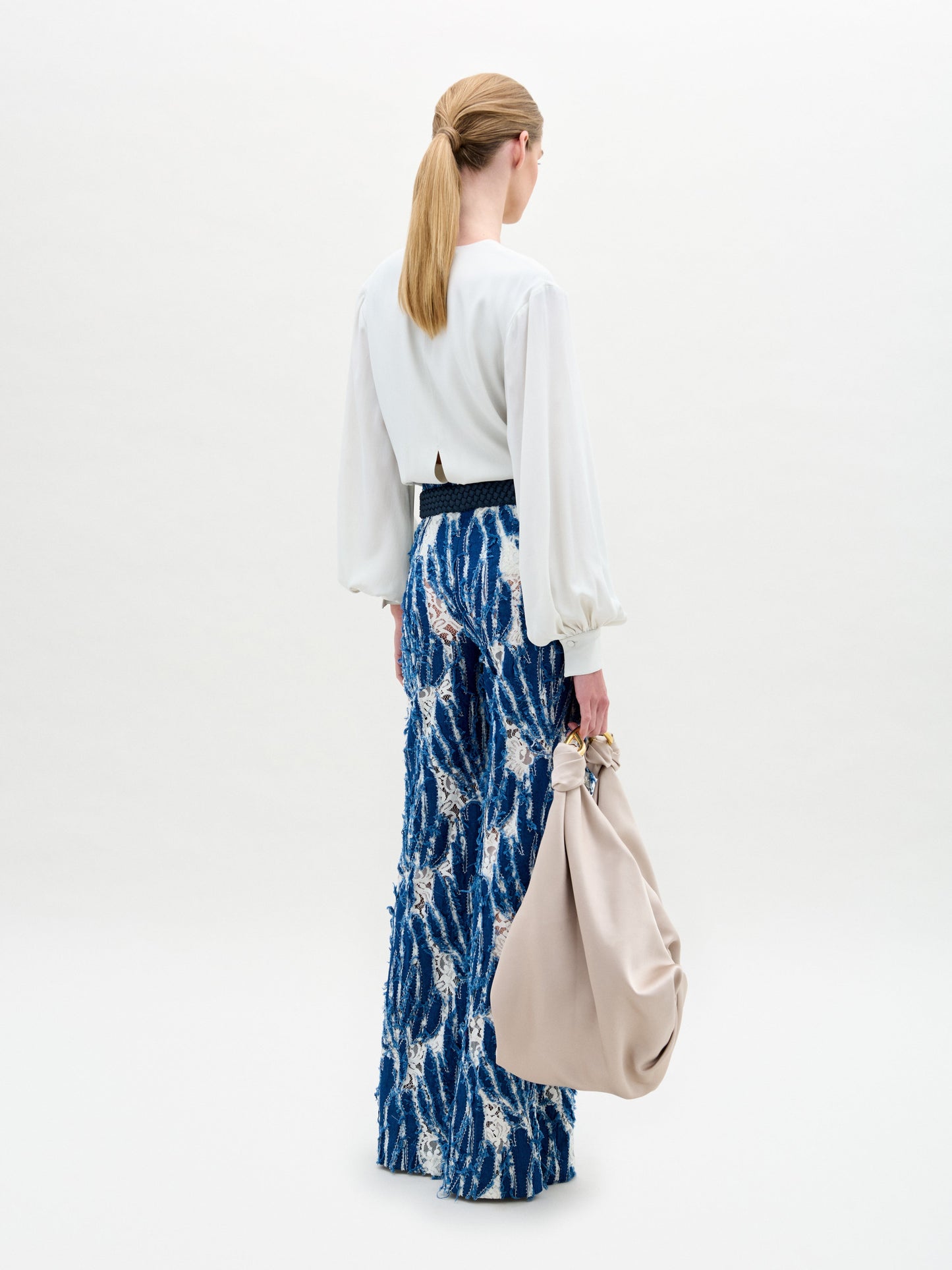 A woman wears the Como Pant, a pair of blue patterned wide-leg pants with a relaxed silhouette, styled with a white long-sleeve front slit blouse and holding a large beige bag against a plain background.