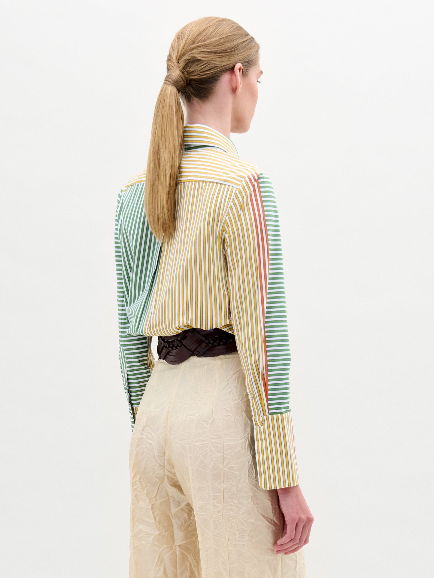 A woman stands against a plain background, wearing the Cubillos Shirt White, a 100% cotton multicolored striped shirt, paired with textured beige pants and a wide belt featuring a large round gold buckle.