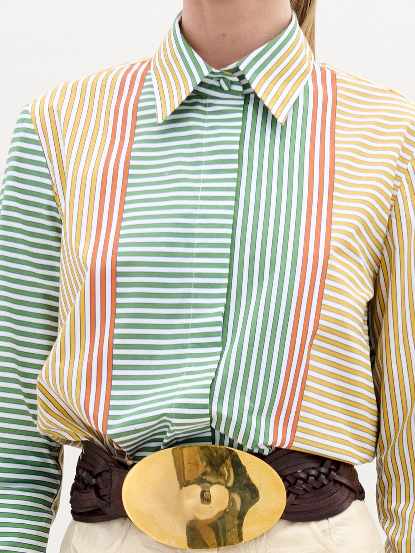 A woman stands against a plain background, wearing the Cubillos Shirt White, a 100% cotton multicolored striped shirt, paired with textured beige pants and a wide belt featuring a large round gold buckle.