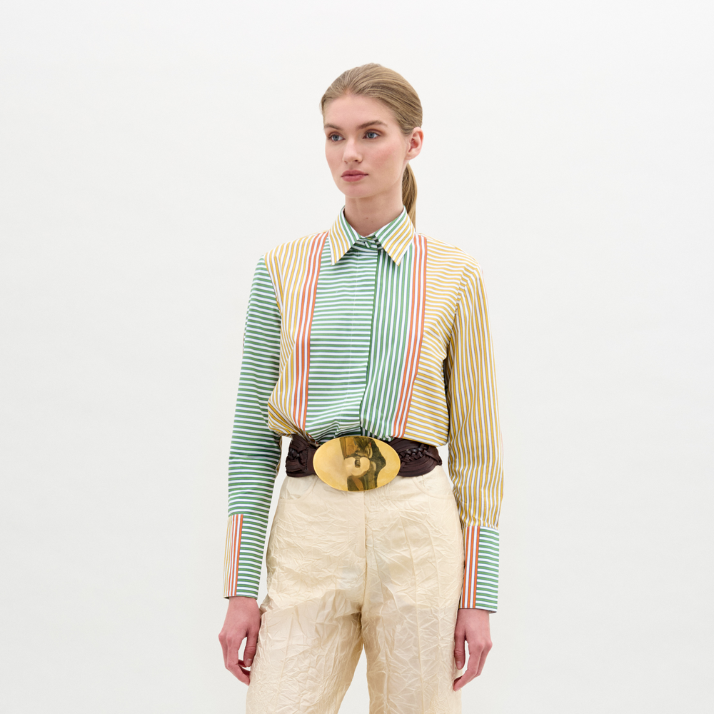 A woman stands against a plain background wearing the Cubillos Shirt, a multicolored 100% cotton striped top, paired with beige textured pants and a large gold belt buckle.