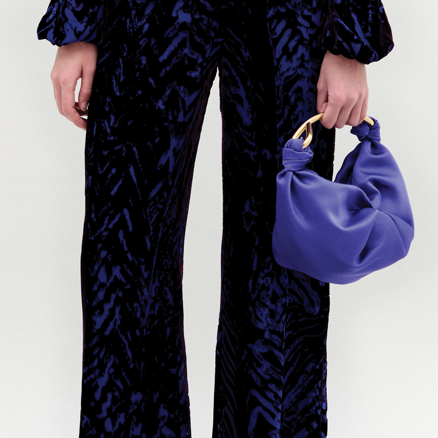 A person stands against a plain background wearing the Como Pant in color eggplant velvet and the sofia bag small in color spectrum blue