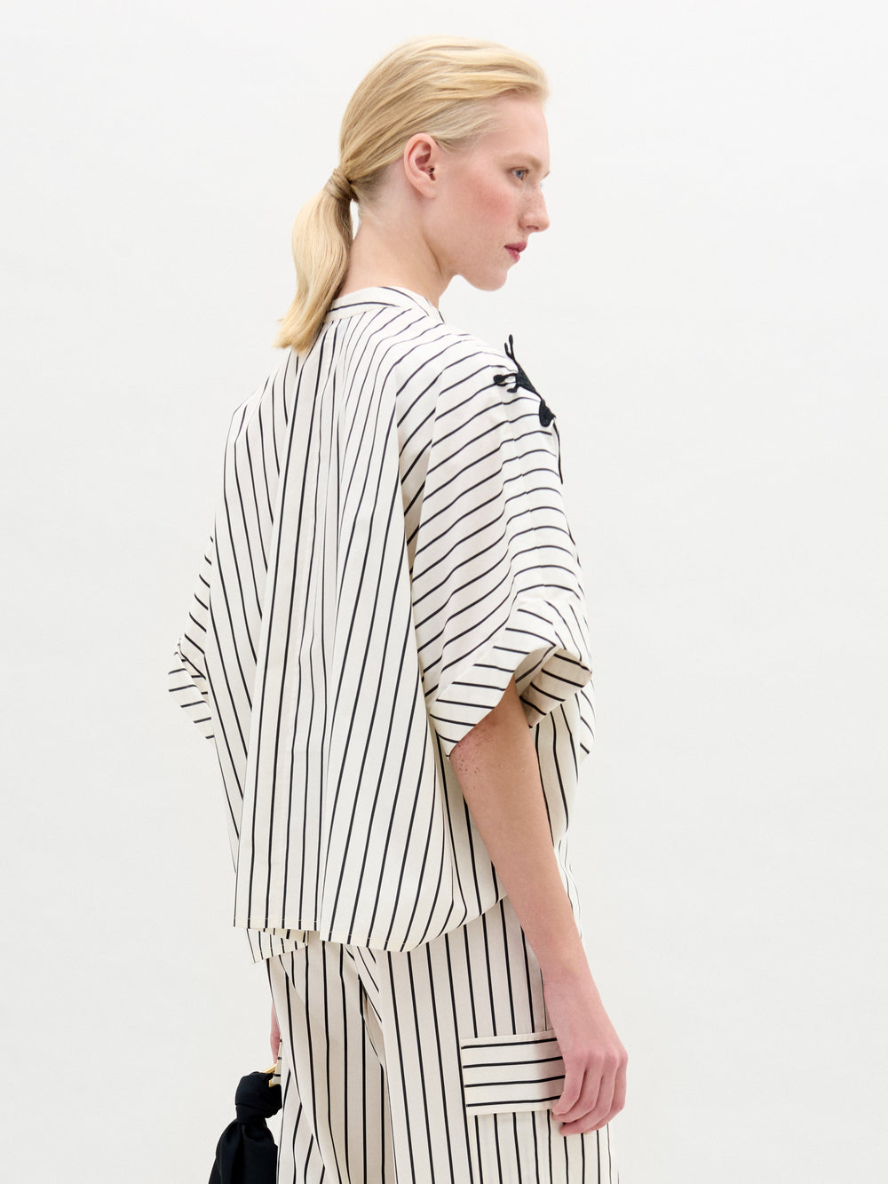 A woman in the Daia Blouse, a white short-sleeved, pinstriped top with a black abstract floral motif on the left shoulder and matching pants in Italian fabric, stands against a plain white background.