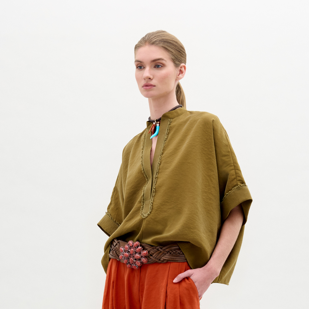 A woman stands against a plain background wearing the olive green Daia Blouse with an oversized fit, orange pants, a woven belt with a floral buckle, and a colorful necklace.