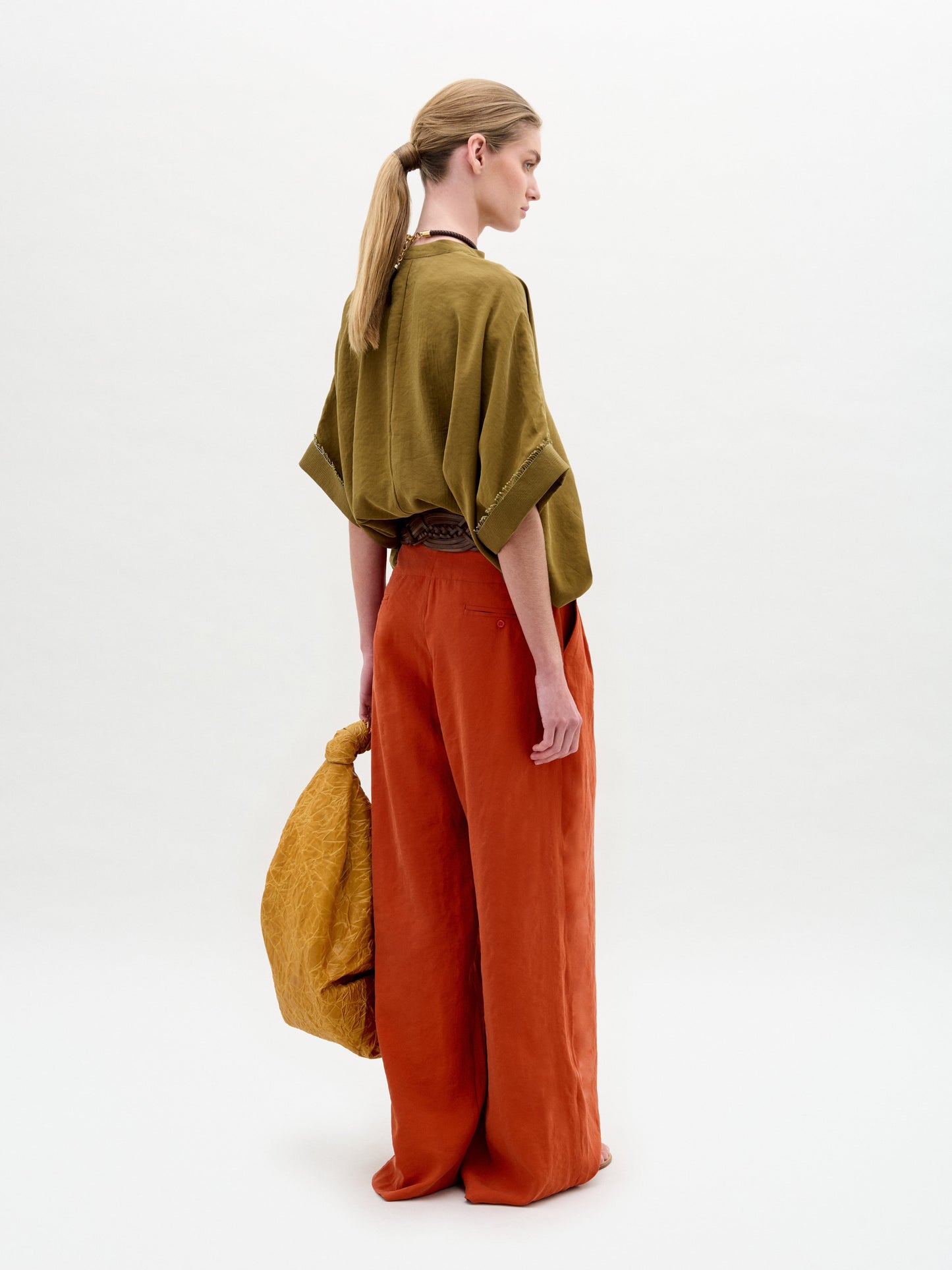 A woman stands sideways against a plain white background, wearing the Daia Blouse in olive green with a V-cut neckline, paired with loose orange pants and holding a textured yellow bag.