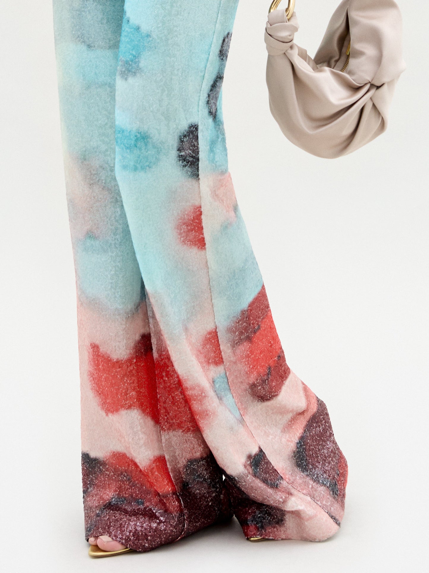 A woman stands against a plain light background wearing the Dalal Pant—high-waisted, wide-leg pants in luxurious Italian fabric with an abstract pattern—styled with a colorful off-the-shoulder top and finished with a yellow belt accent.