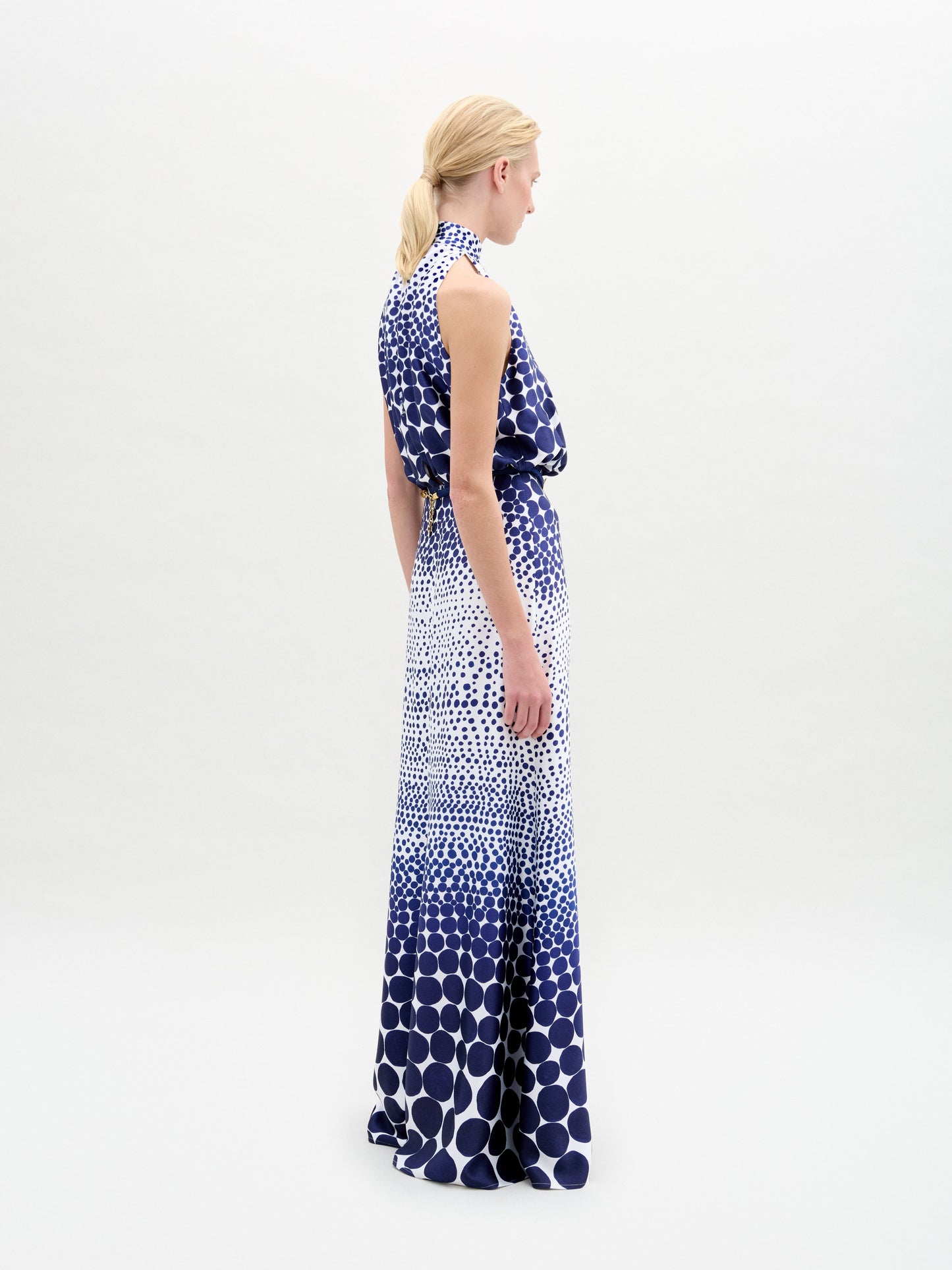 A person wears the Damara Dress, a sleeveless, high-neck blue and white dotted maxi, cinched with a gold-buckled 3D Cord belt, standing against a plain white background.