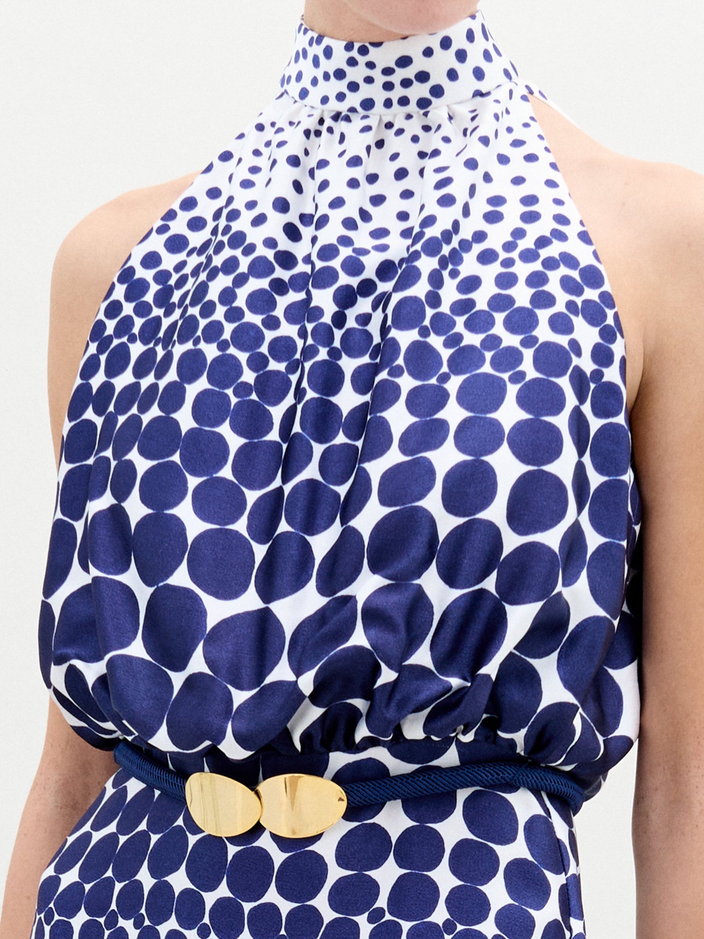 A person wears the Damara Dress, a sleeveless, high-neck blue and white dotted maxi, cinched with a gold-buckled 3D Cord belt, standing against a plain white background.