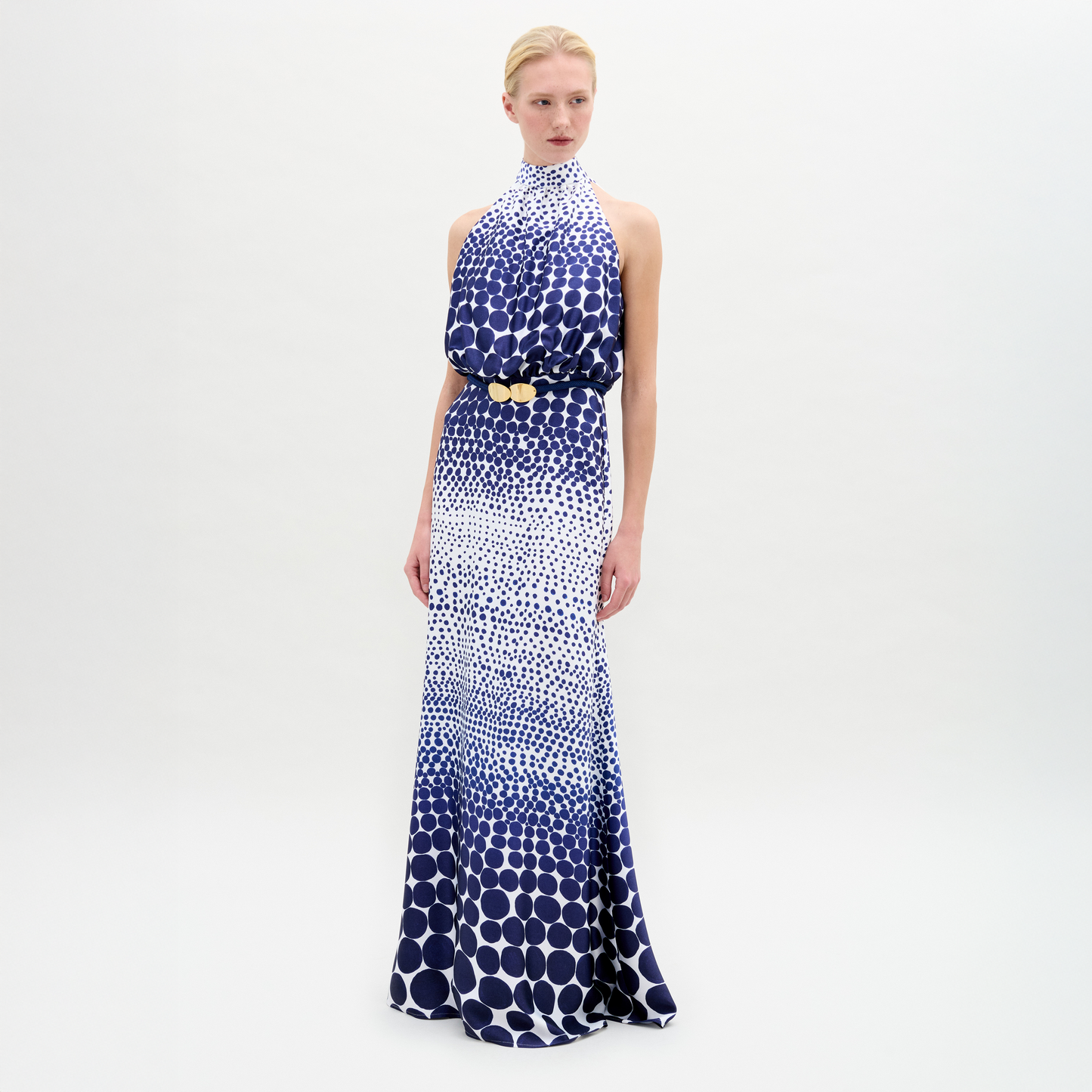 A person wears the Damara Dress, a sleeveless, high-neck blue and white dotted maxi, cinched with a gold-buckled 3D Cord belt, standing against a plain white background.