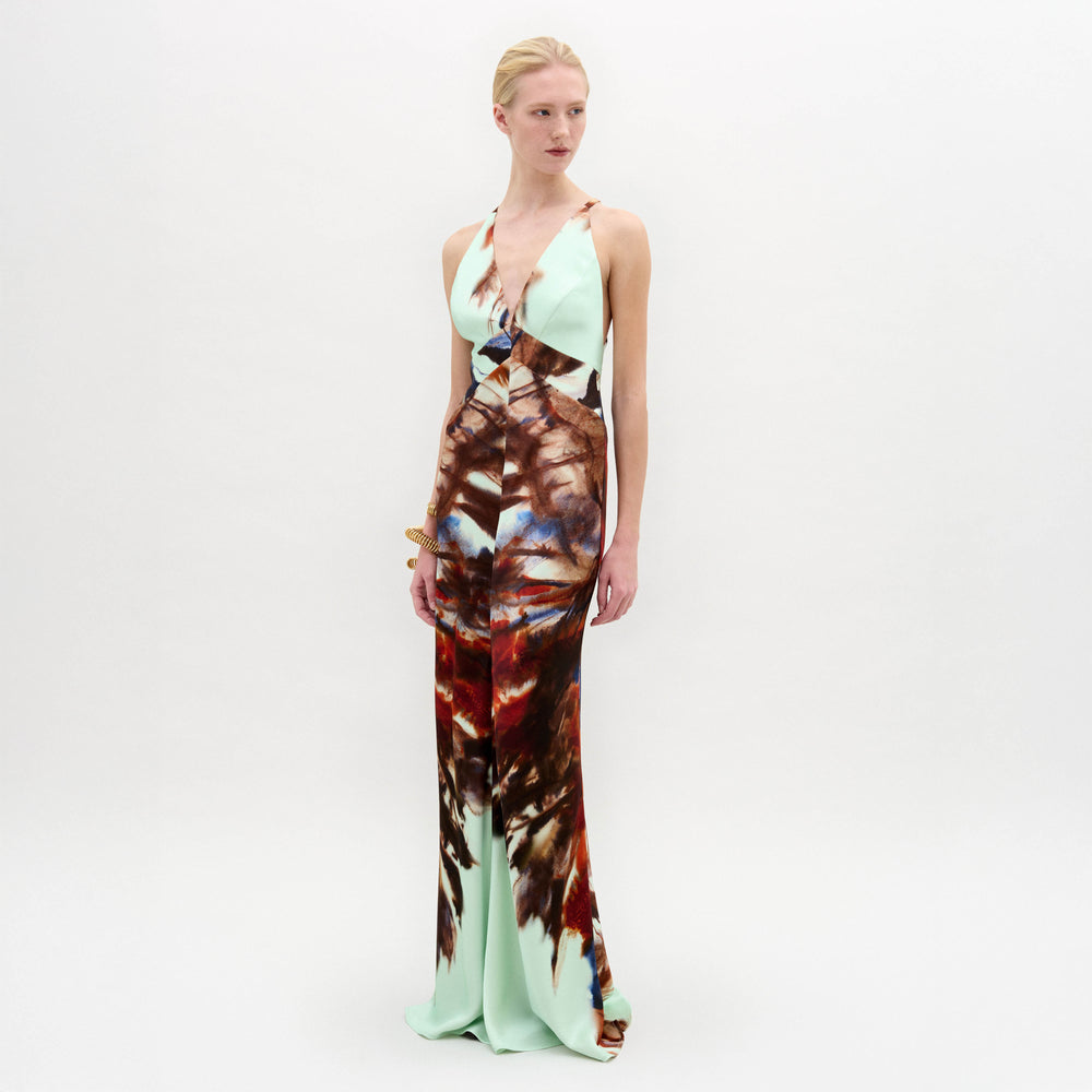 A woman stands against a plain white background wearing the Daniella Dress—a sleeveless maxi with a deep V-neckline, featuring a light blue top and a brown and red abstract print on the lower half.