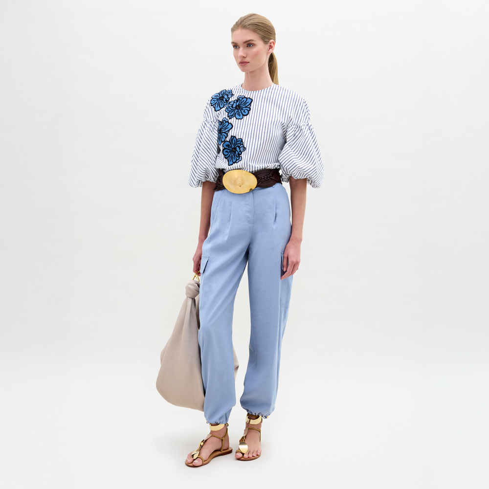 A woman stands against a plain background wearing a striped blouse with blue floral appliqué, the Darnell Pant in light blue, a brown belt with a large buckle, gold sandals, and holding a beige bag.