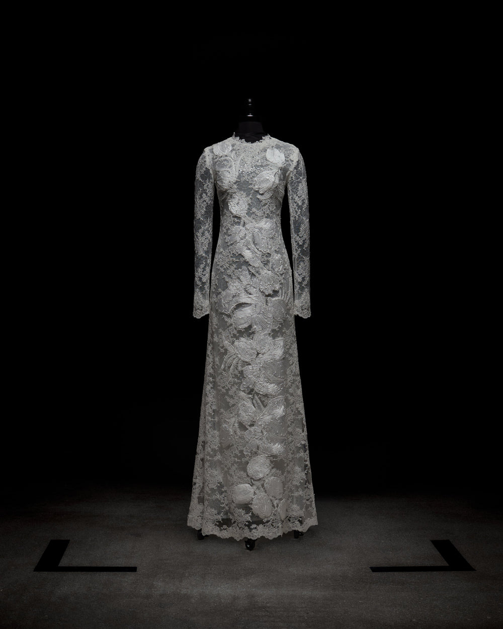 A person stands against a plain white background, arms crossed, wearing the Dava Dress—a sheer, long-sleeved lace dress with floral patterns.