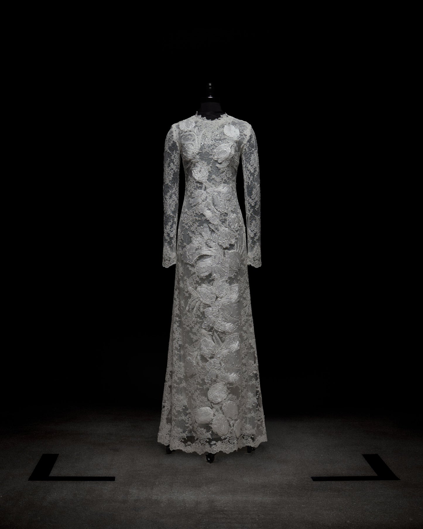 A person stands against a plain white background, arms crossed, wearing the Dava Dress—a sheer, long-sleeved lace dress with floral patterns.