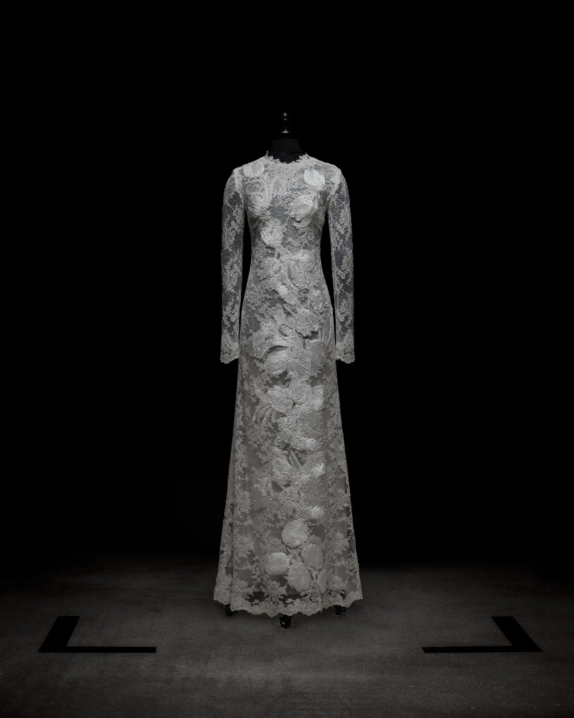A person stands against a plain white background, arms crossed, wearing the Dava Dress—a sheer, long-sleeved lace dress with floral patterns.