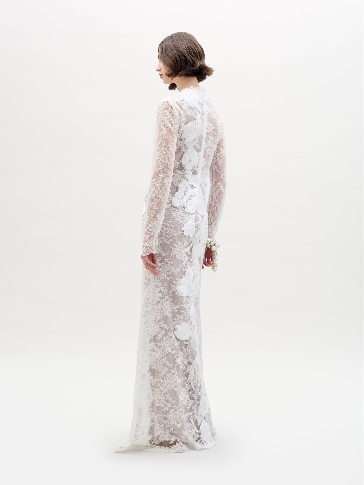 A person stands against a plain white background, arms crossed, wearing the Dava Dress—a sheer, long-sleeved lace dress with floral patterns.