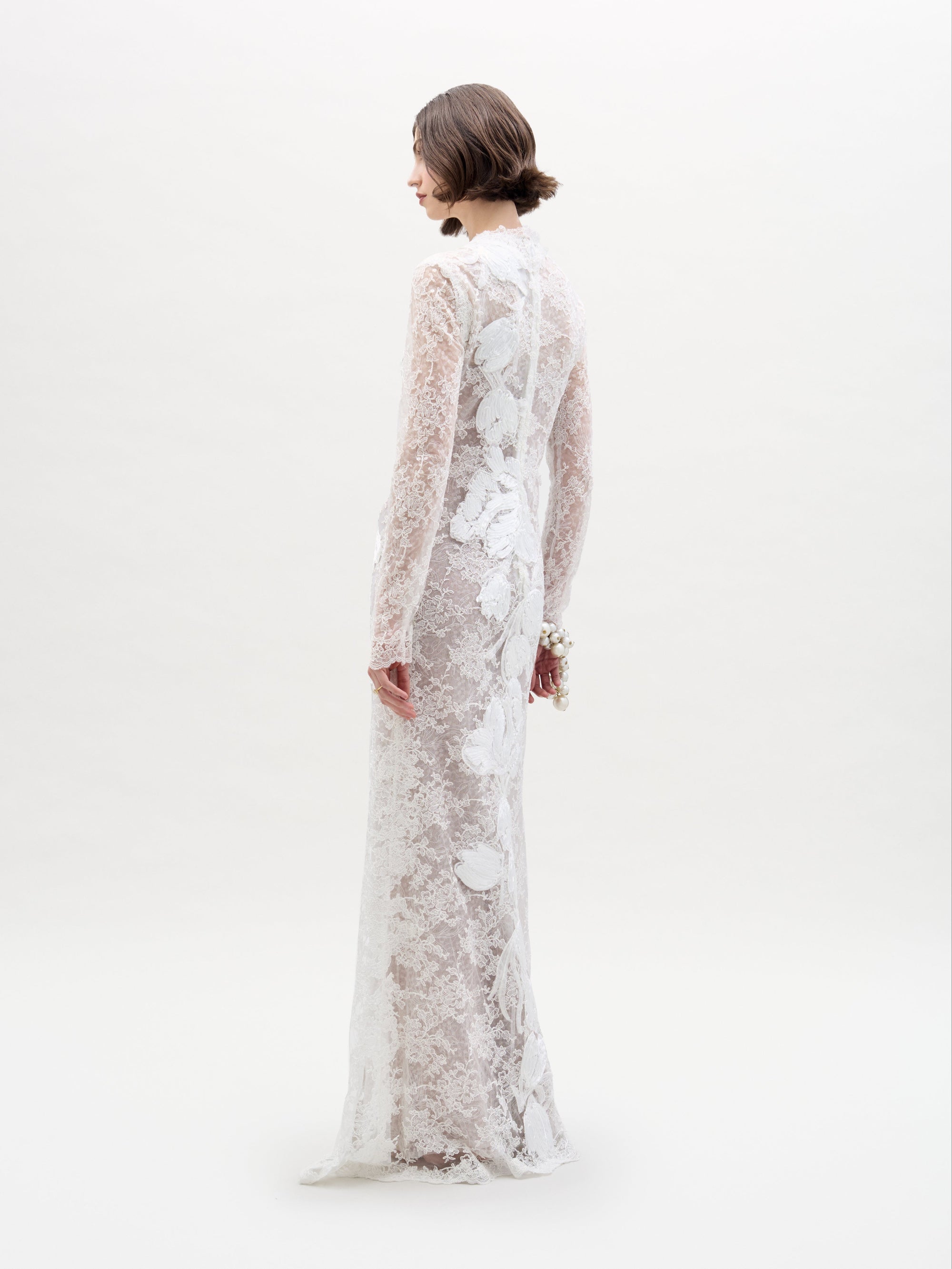 A person stands against a plain white background, arms crossed, wearing the Dava Dress—a sheer, long-sleeved lace dress with floral patterns.