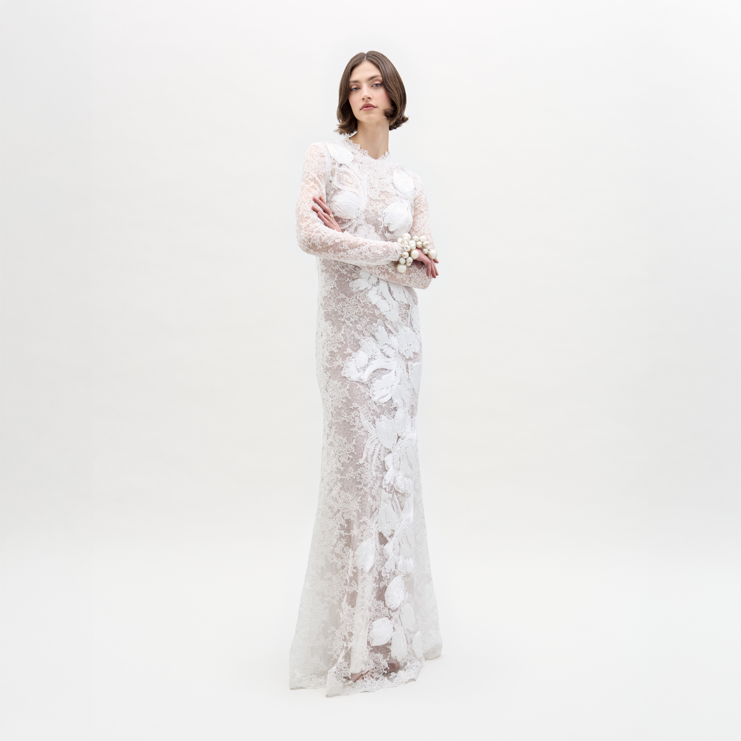 A person stands against a plain white background, arms crossed, wearing the Dava Dress—a sheer, long-sleeved lace dress with floral patterns.