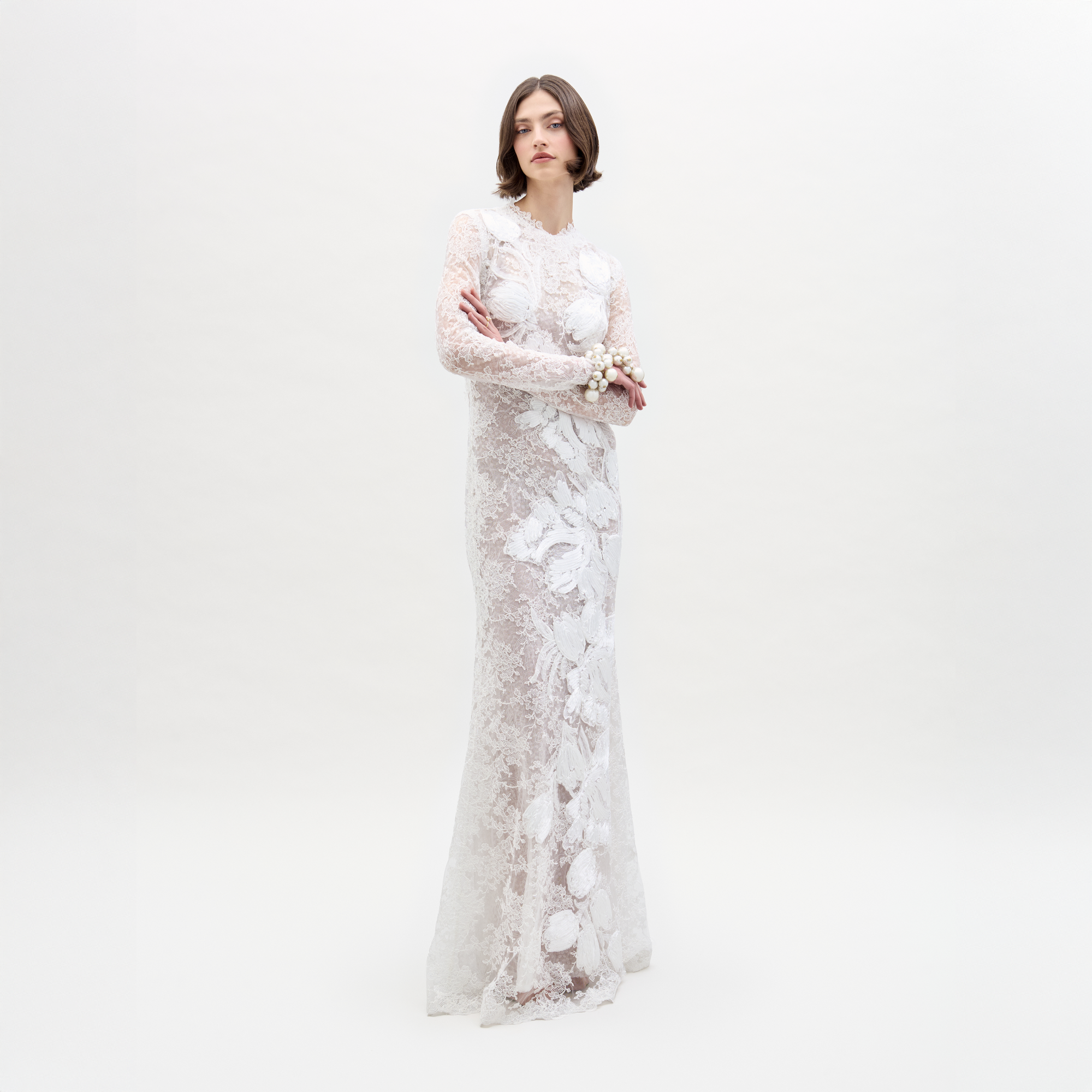 A person stands against a plain white background, arms crossed, wearing the Dava Dress—a sheer, long-sleeved lace dress with floral patterns.