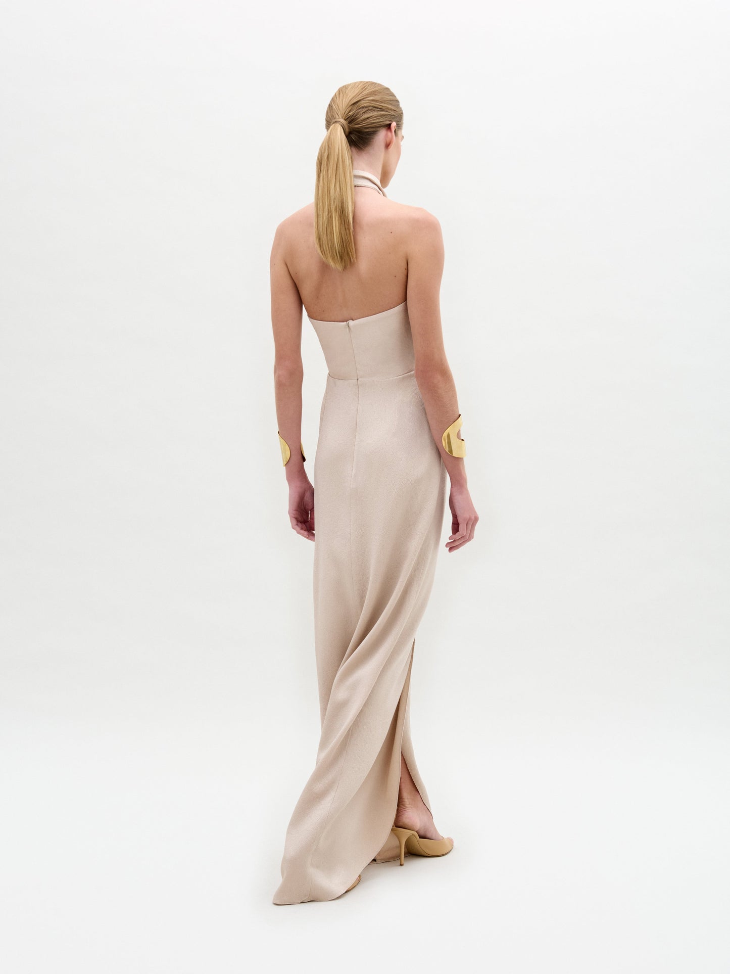 A woman wears the Davinia Dress, a cream, floor-length halter gown with a floral accent at the waist, paired with a gold cuff bracelet and beige shoes, standing against a plain white background.
